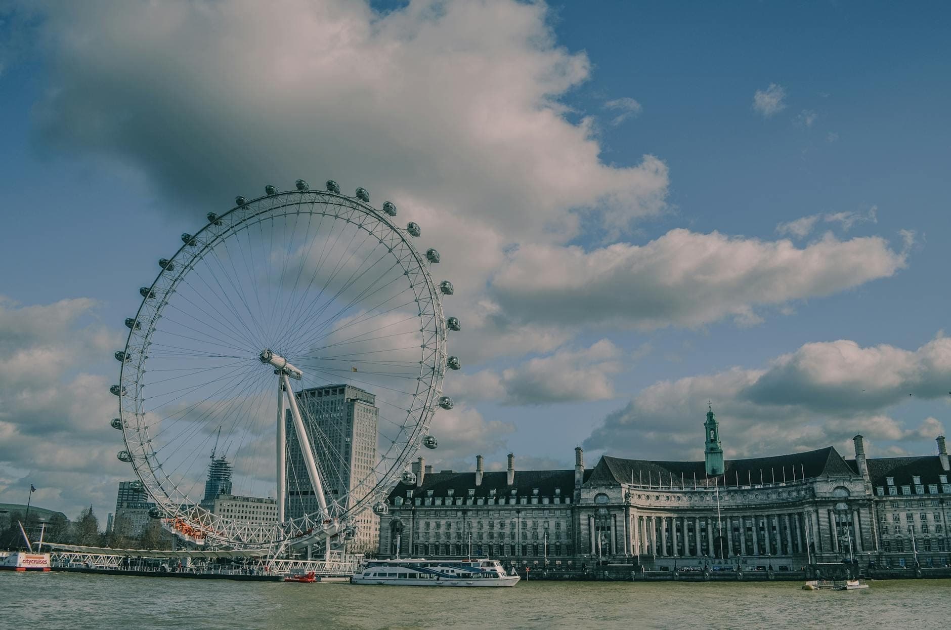 La noria del London Eye dominando el Támesis, con el County Hall y barcos en el río bajo un cielo azul brillante con nubes esponjosas.