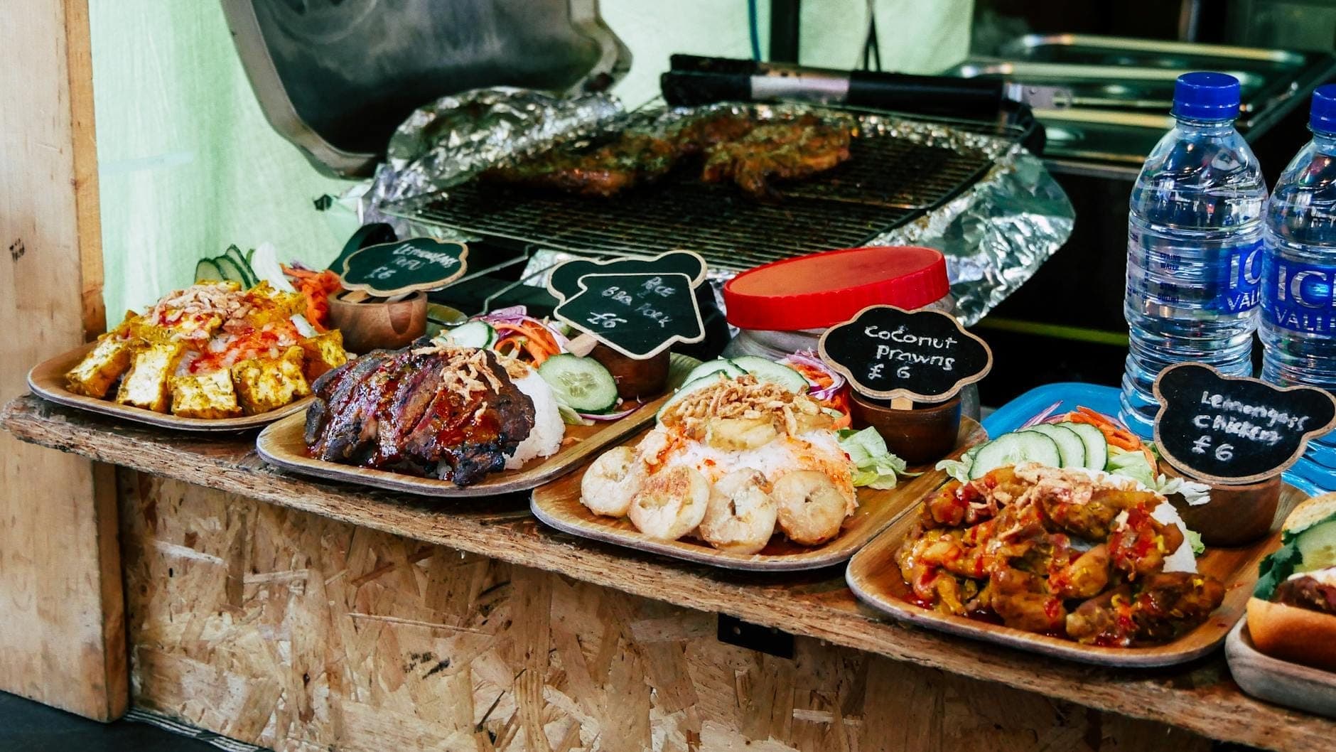 Kolorowa ekspozycja różnorodnych street foodów na londyńskim targu — grillowane mięsa, krewetki, kurczak, sałatki, sosy i etykiety z cenami w funtach.