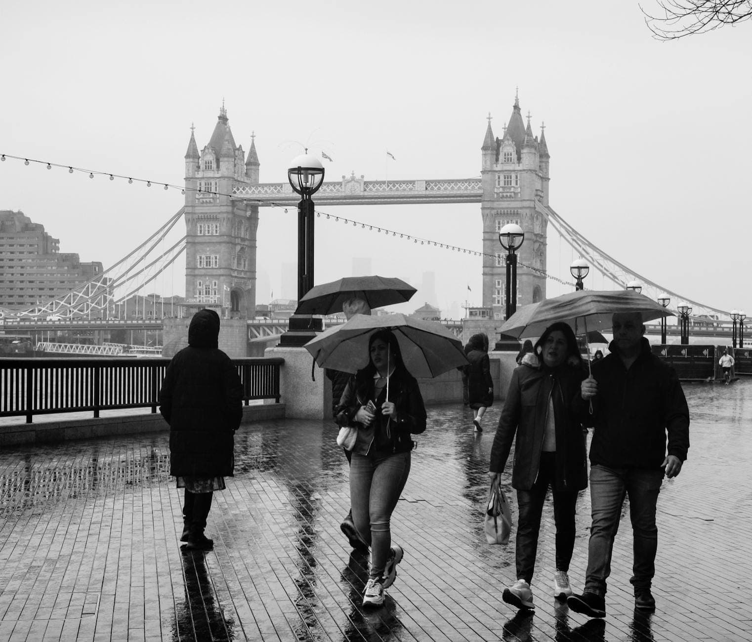 Personas caminando con paraguas en un día lluvioso cerca del Tower Bridge en Londres, pavimento mojado y cielo nublado.