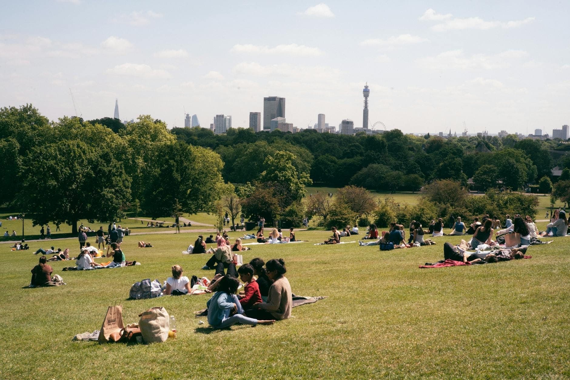 Ludzie odpoczywający na trawiastym wzgórzu z panoramicznym widokiem na panoramę Londynu i słynne zabytki w oddali.