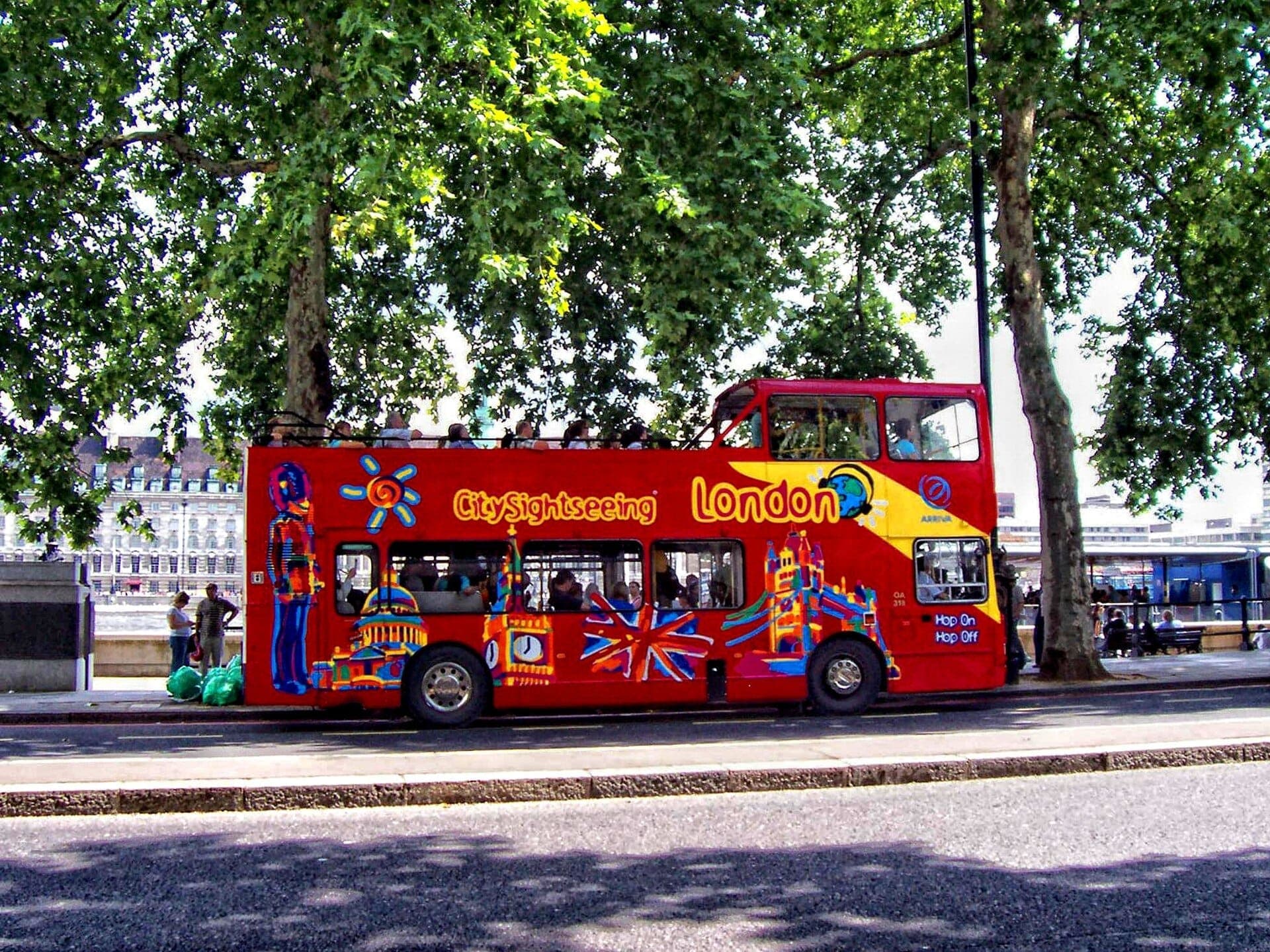 Leuchtend roter City Sightseeing London Doppeldecker-Hop-on-Hop-off-Bus unter belaubten Bäumen geparkt, mit ikonischen Stadtsehenswürdigkeiten und dem Fluss im Hintergrund.