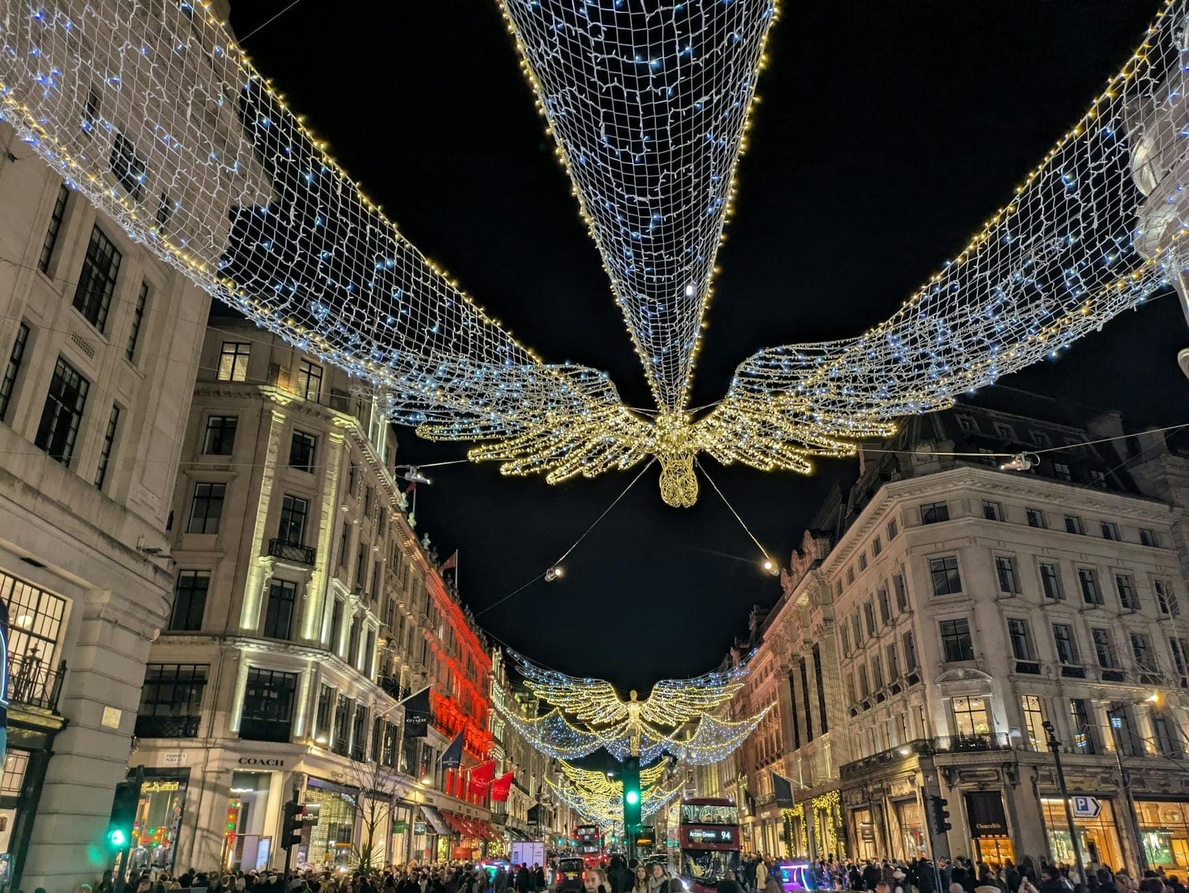 Tłumy podziwiają mieniące się świąteczne światełka w kształcie aniołów rozwieszone nad ruchliwą ulicą w centrum Londynu nocą.