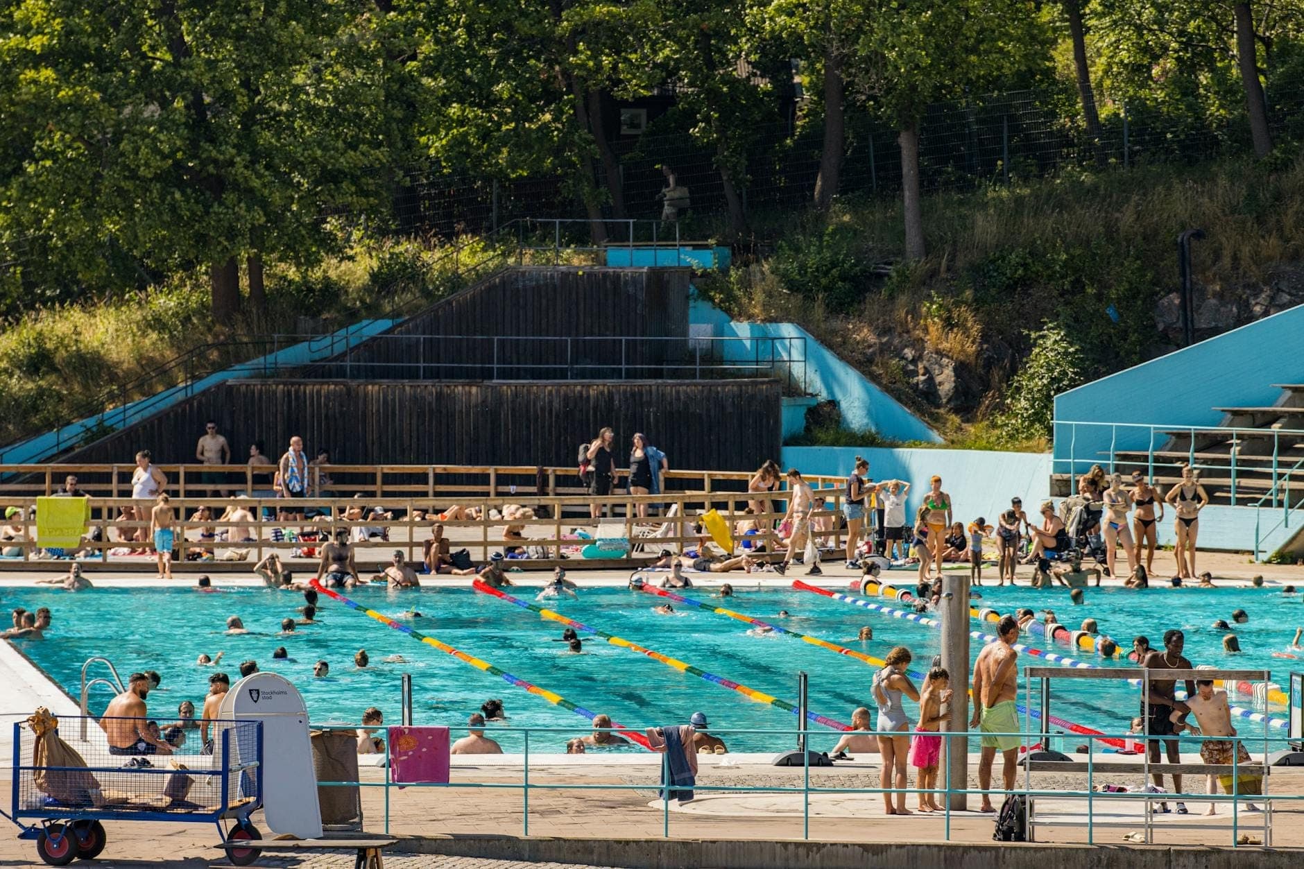 Una concurrida piscina pública al aire libre con multitudes de personas disfrutando del sol, rodeada de árboles y paredes azules, en un día de verano.