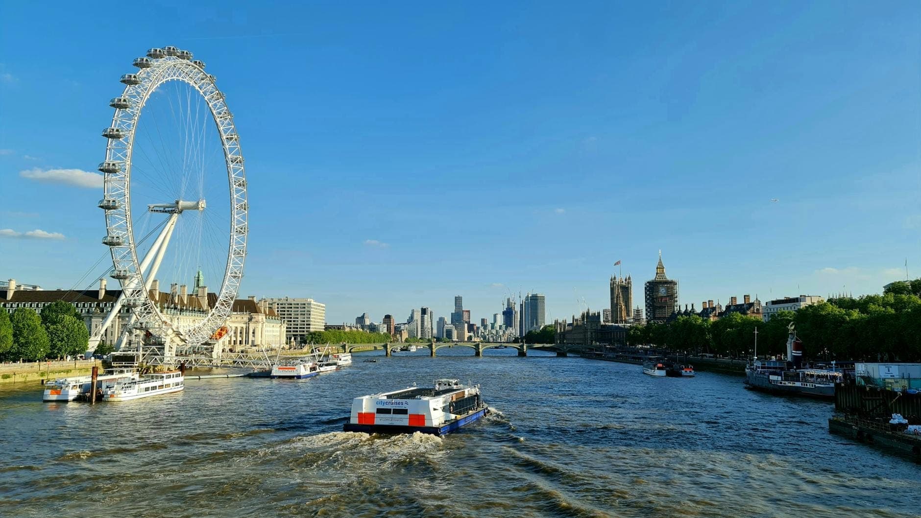 Vista panorámica del río Támesis en un día soleado con el London Eye, embarcaciones en el agua y el horizonte de la ciudad al fondo.
