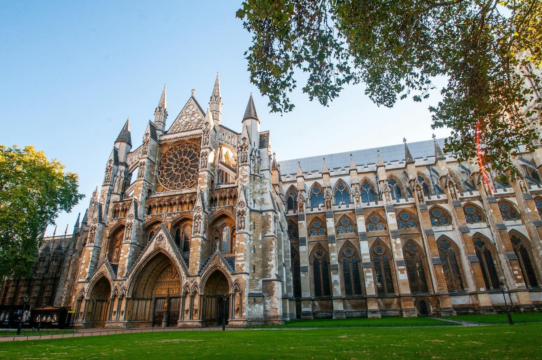 Weitwinkelansicht der Hauptfassade und des Rasens der Westminster Abbey, mit gotischen Bögen, großem Rosettenfenster und Bäumen im Hintergrund.