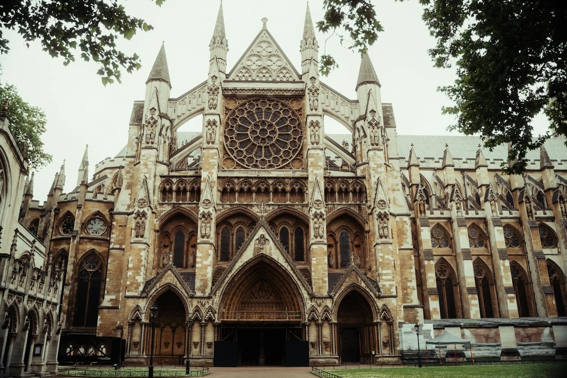Vista frontale completa dell'esterno della Westminster Abbey con intricata lavorazione in pietra, rosone e torri appuntite, incorniciata da alberi frondosi su entrambi i lati.