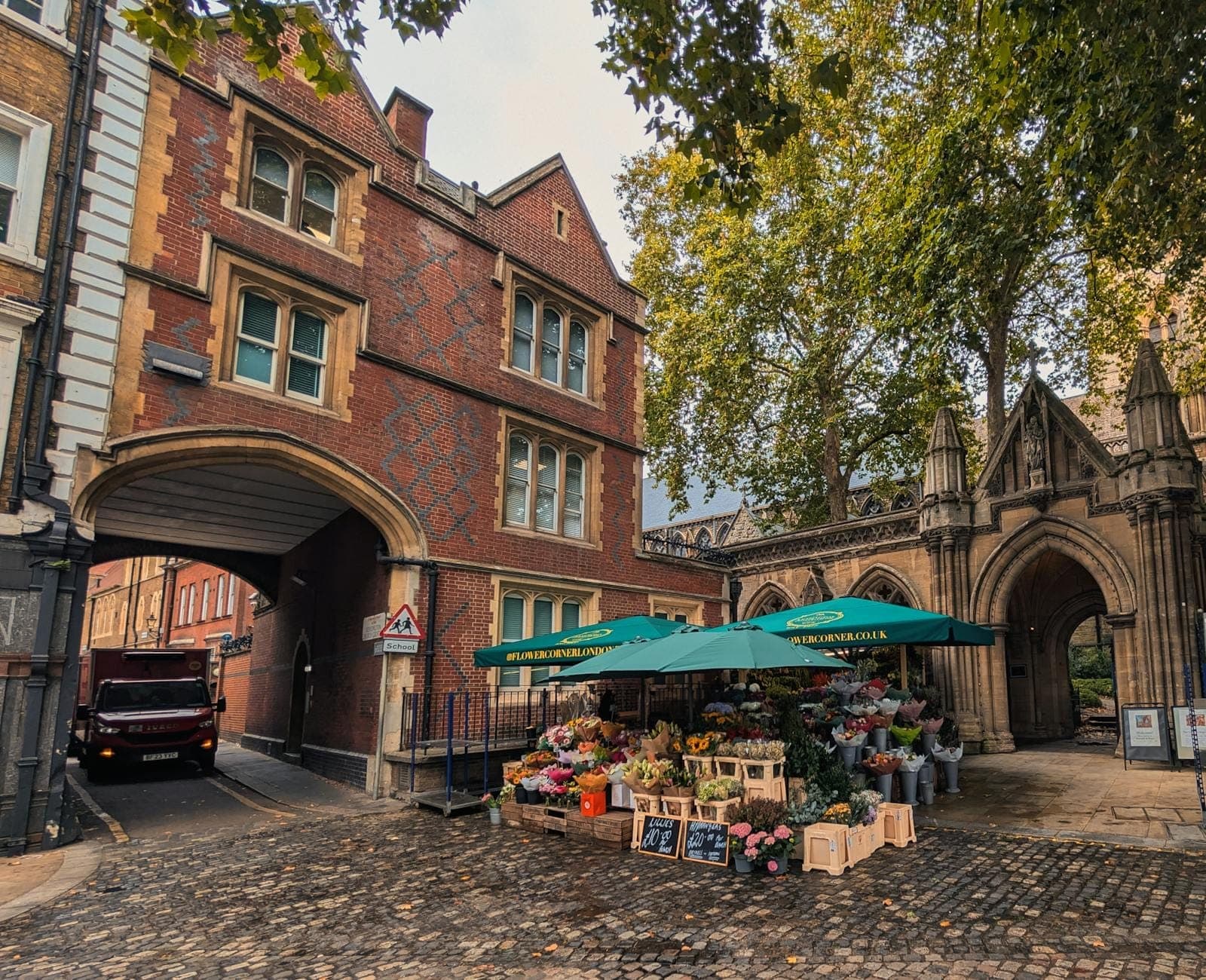Blumenmarktstand unter grünen Schirmen vor historischen roten Backsteingebäuden und einem steinernen Torbogen auf einer Kopfsteinpflasterstraße in London.