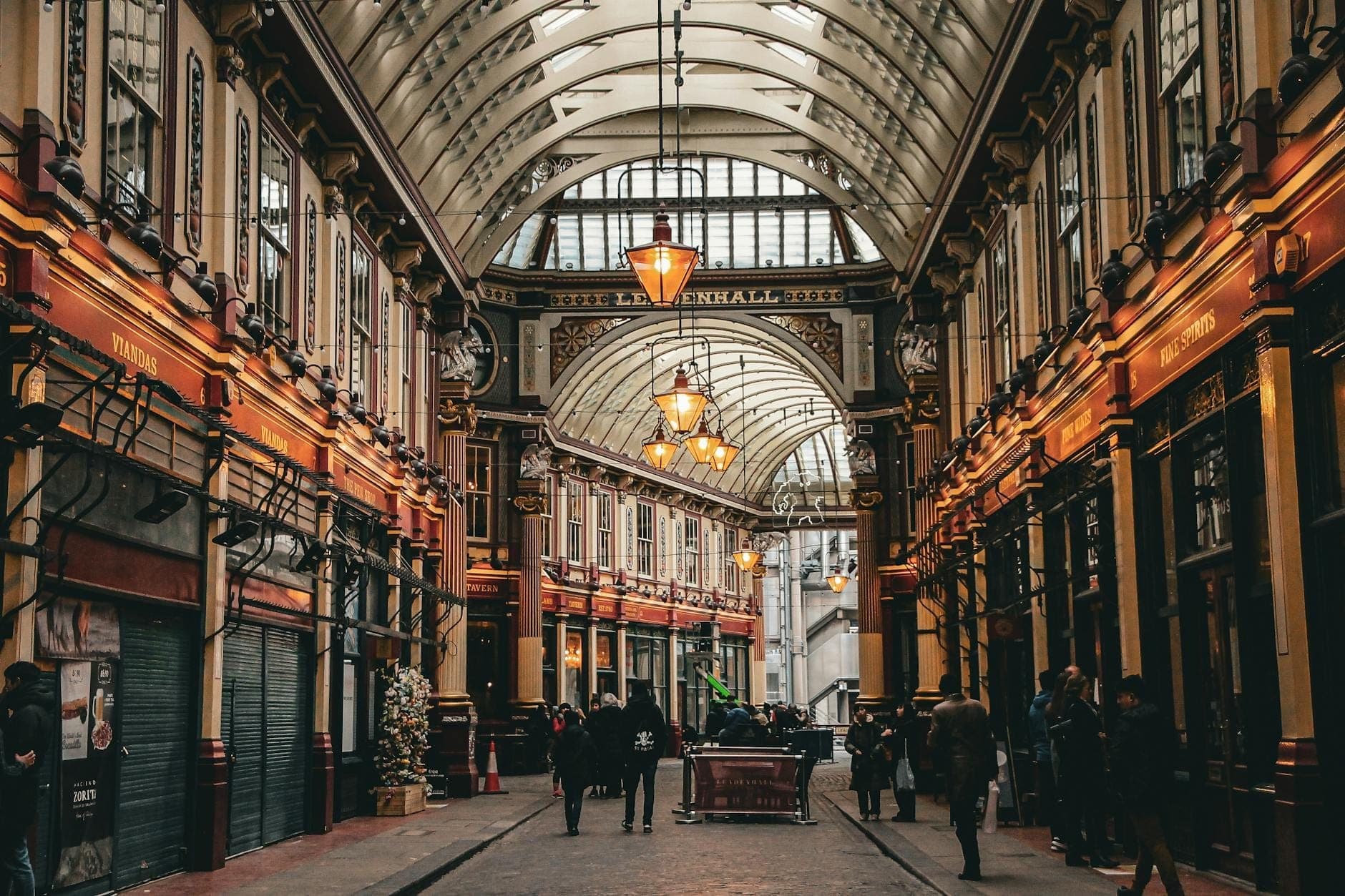 Weitwinklige Ansicht des verzierten, glasüberdachten Innenraums des historischen Leadenhall Market in London, mit Menschen, die unter viktorianischer Architektur und warmem Licht spazieren.