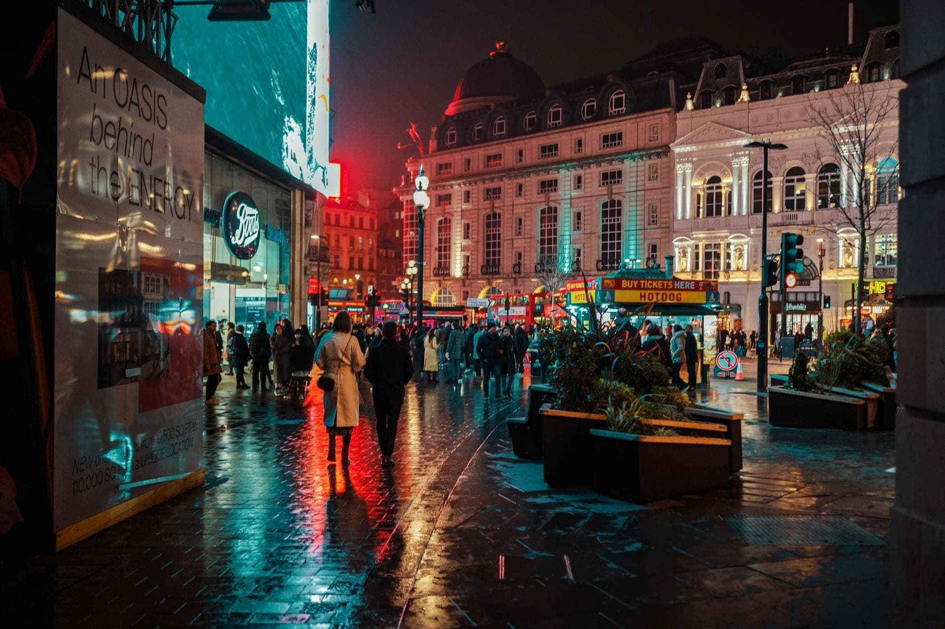 Tłoczna ulica w centrum Londynu nocą z tłumami, neonami i odbiciami świateł, oddająca atmosferę tętniącego życiem miasta.