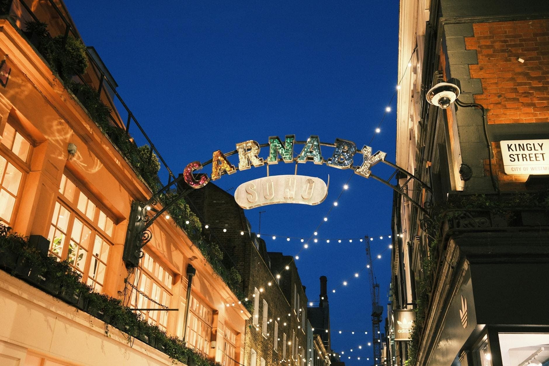 Wejście na Carnaby Street rozświetlone nocą festiwnymi światłami, otoczone ikonicznymi budynkami Londynu, ukazujące słynną dzielnicę życia nocnego.
