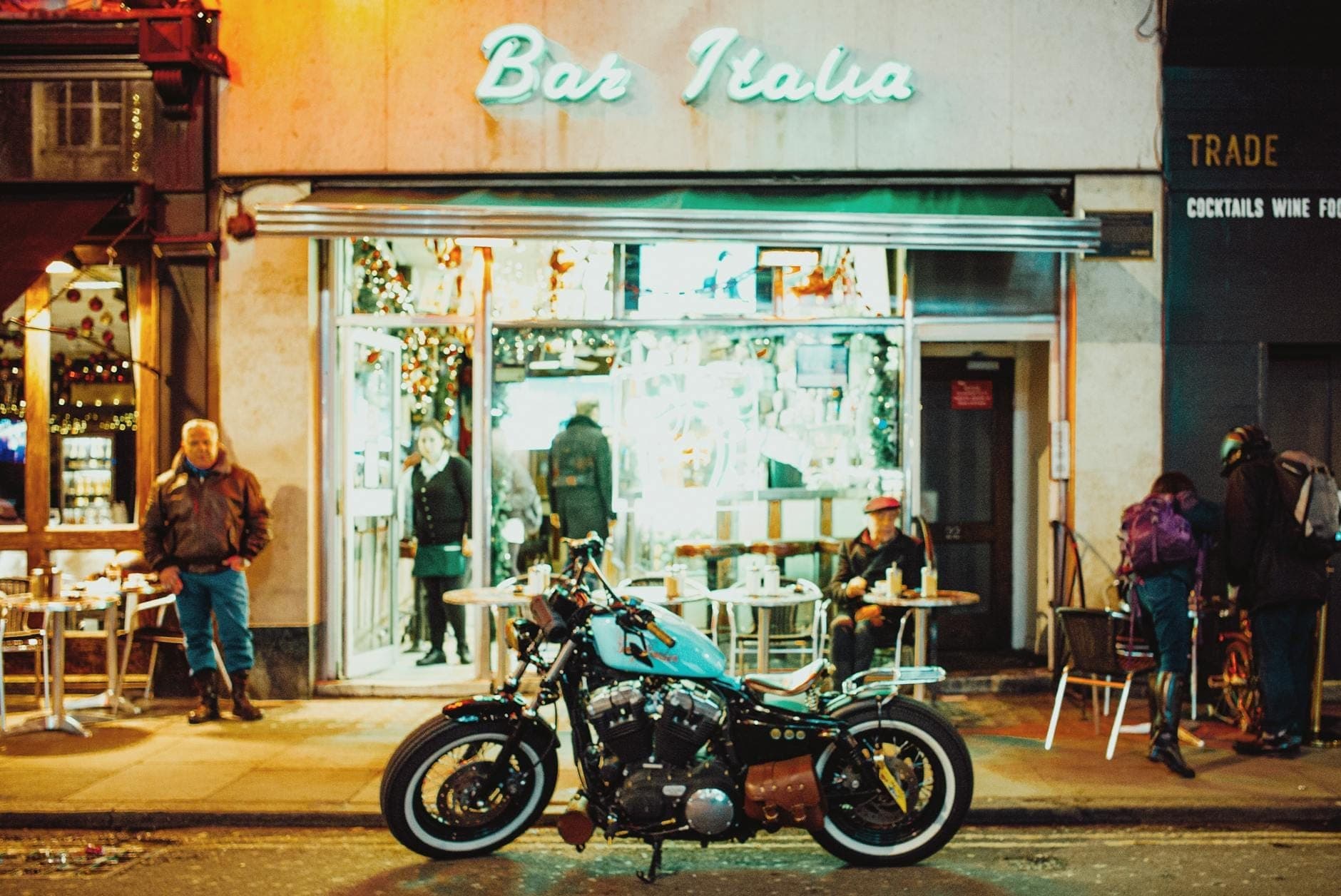 Widok ulicy Bar Italia w Londynie nocą z ludźmi siedzącymi na zewnątrz i zaparkowanym przed wejściem motocyklem.