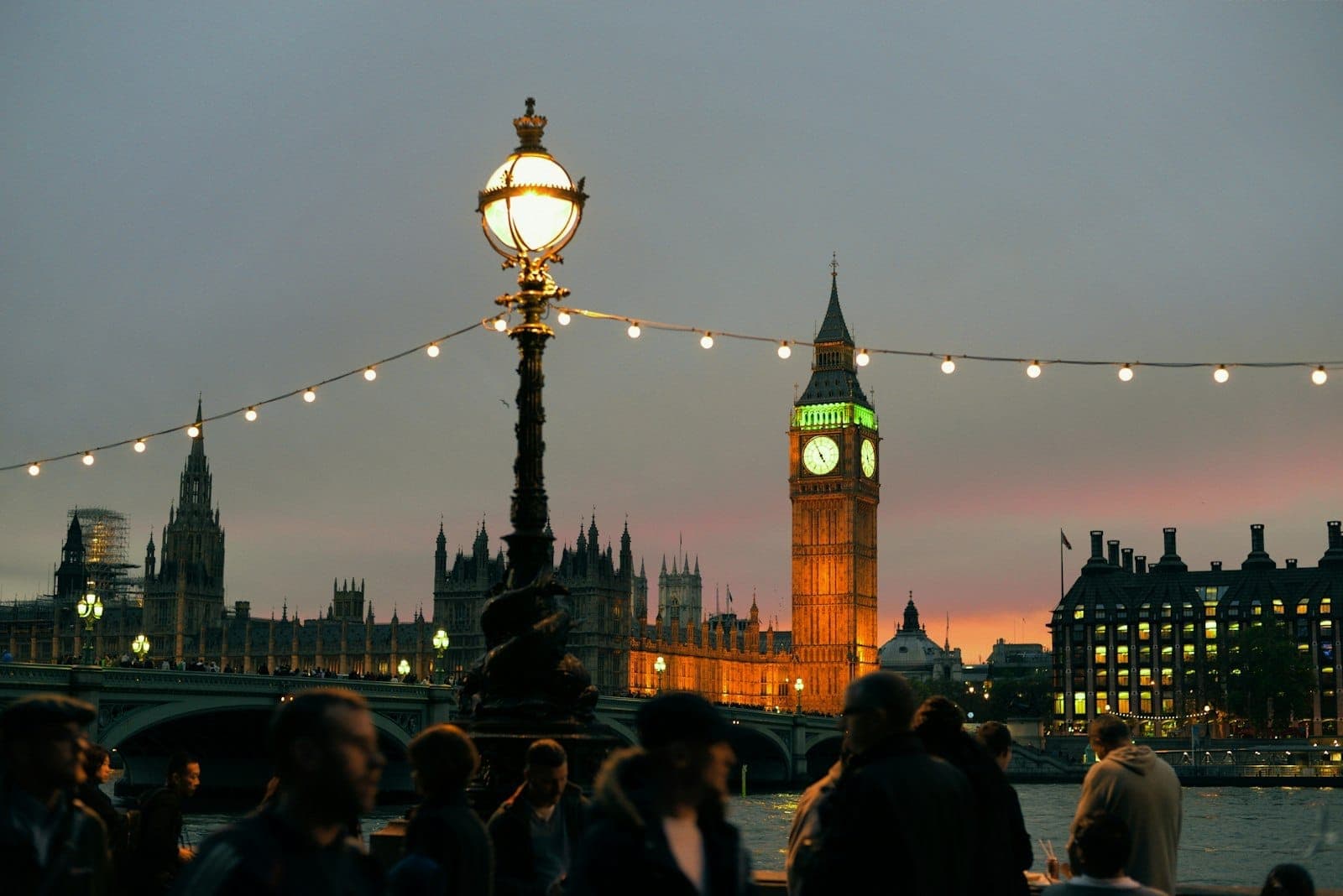Vista noturna de Big Ben e do Parlamento com postes de luz, cordões de luzes e pessoas caminhando ao longo do Tâmisa em Londres.
