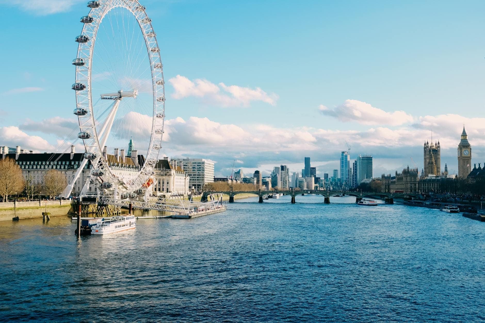 London Eye, Tamiza i Big Ben pod bezchmurnym błękitnym niebem z łodziami i miejskimi budynkami widocznymi wzdłuż wody.