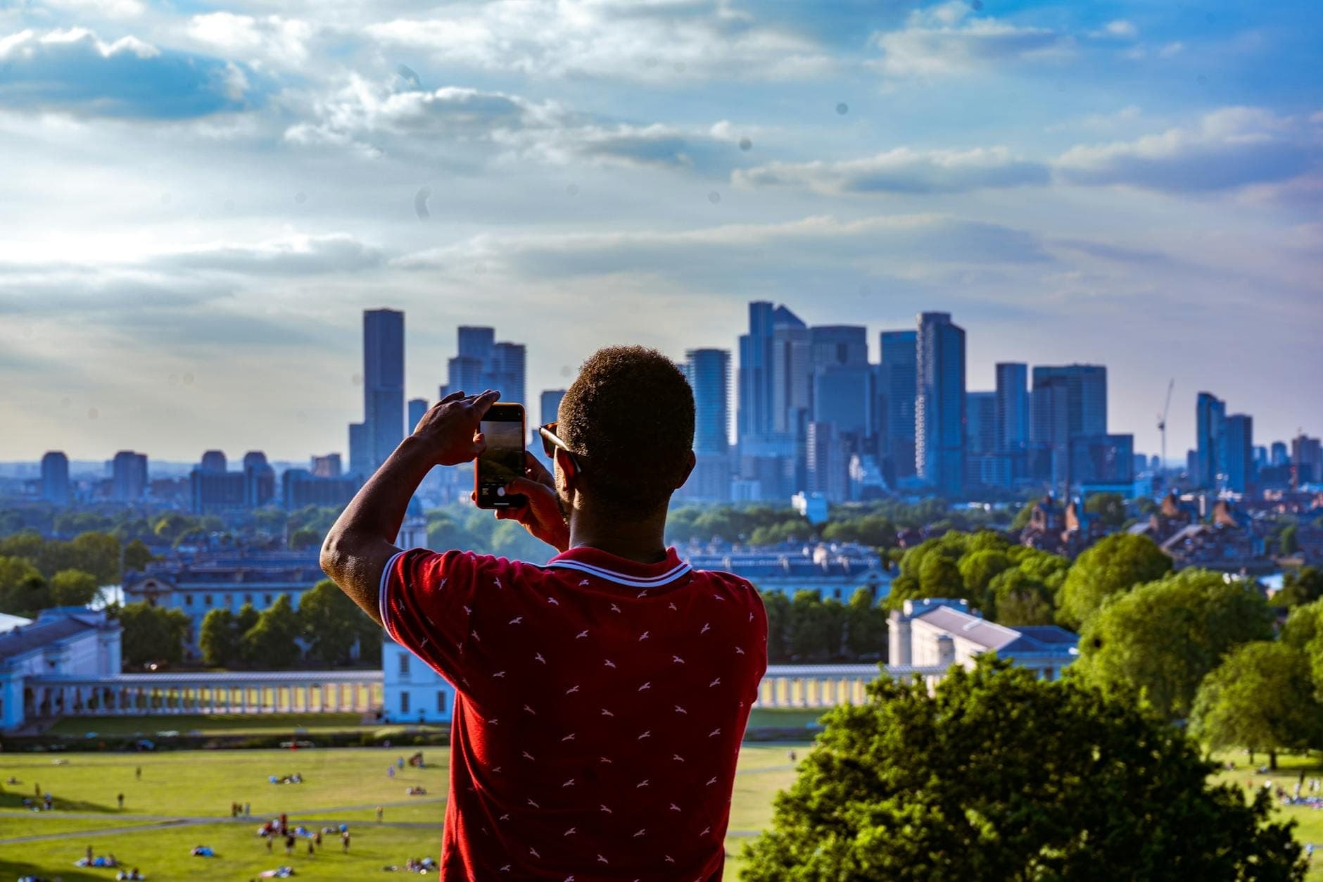 Osoba fotografująca panoramę Londynu z Greenwich Park w słoneczny dzień, z widocznymi poniżej zabytkami miasta i zielonymi trawnikami.