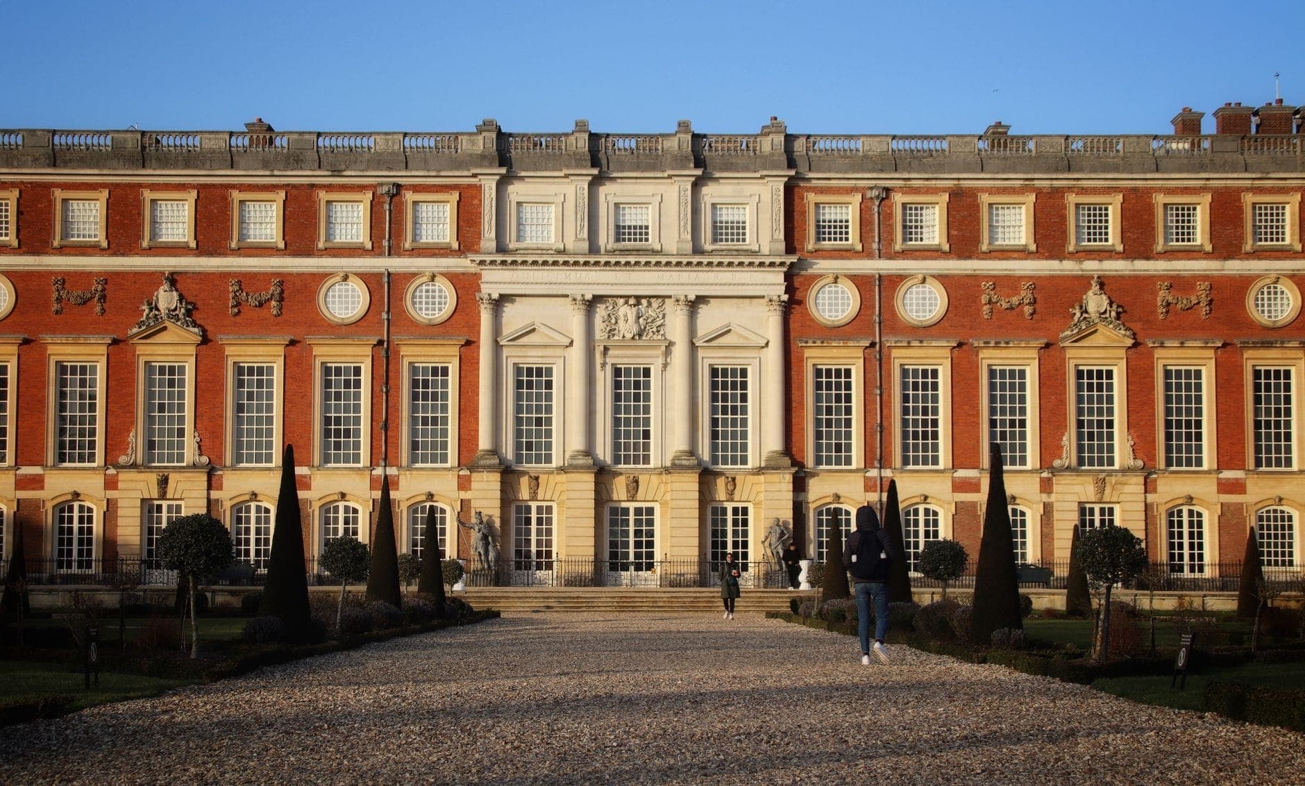 Szeroki, słoneczny widok na Hampton Court Palace z ludźmi spacerującymi po historycznej żwirowej ścieżce, okazałą symetryczną fasadą i formalnymi ogrodami na pierwszym planie.
