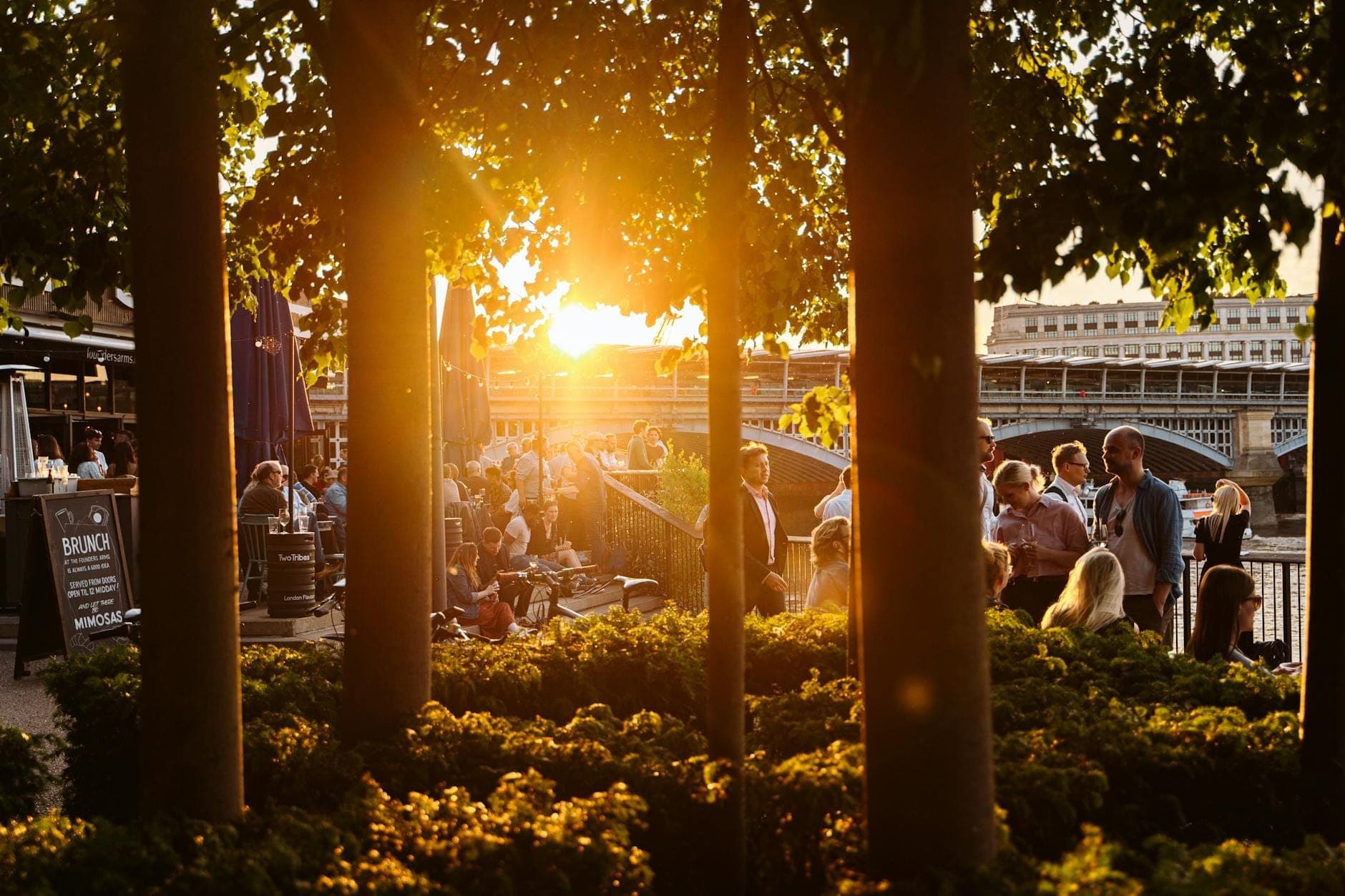 People enjoying food and drinks at outdoor riverside venues along the Thames with sunset and greenery in central London.