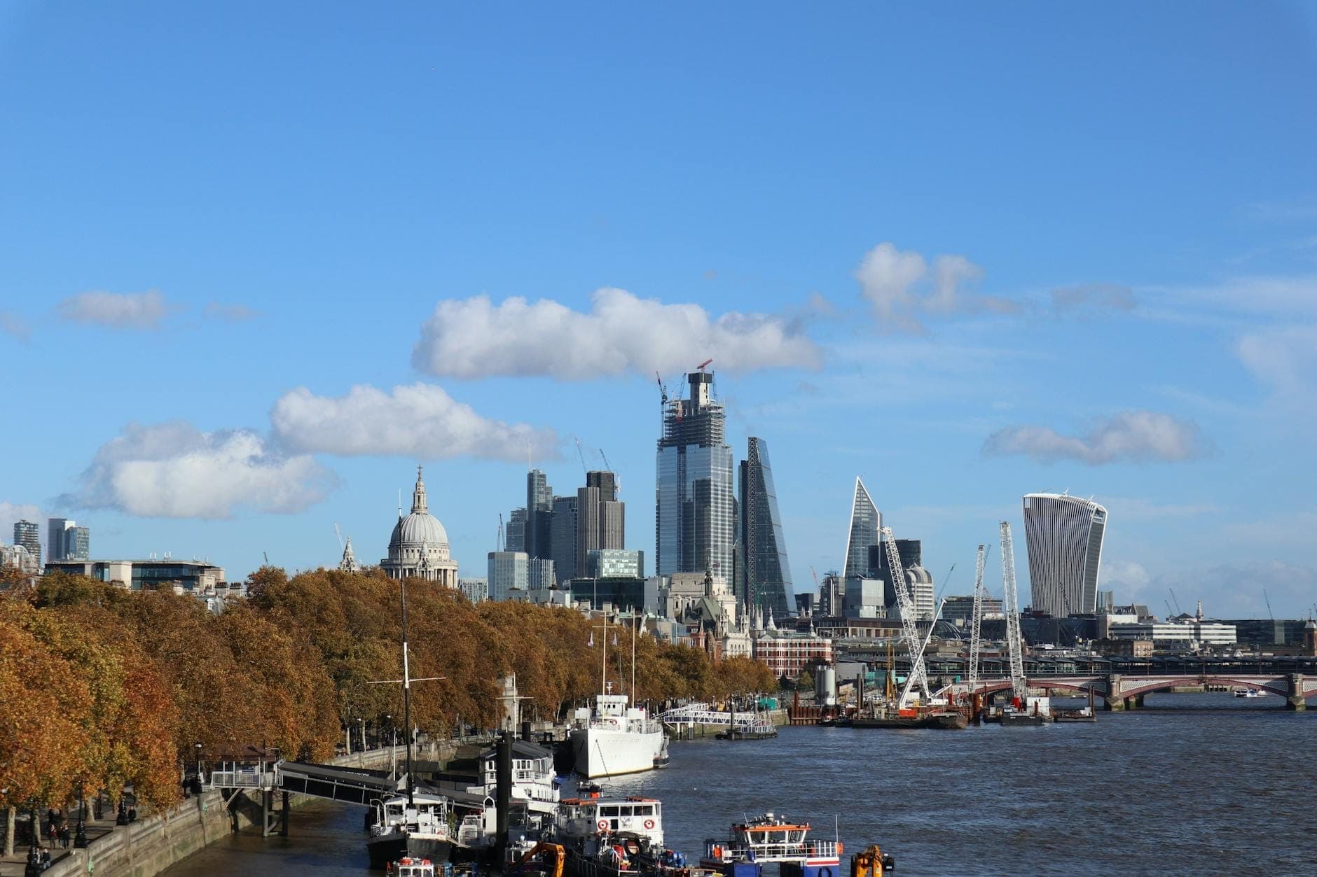 View of the River Thames in London with boats, autumn trees, and modern city skyline under a clear blue sky.