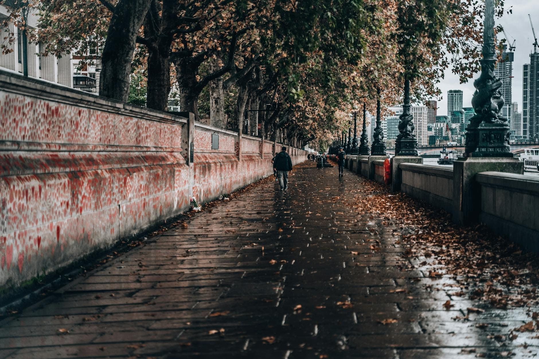 Riverside path in London with autumn leaves, people walking, benches, and distant city skyline under tree canopy.