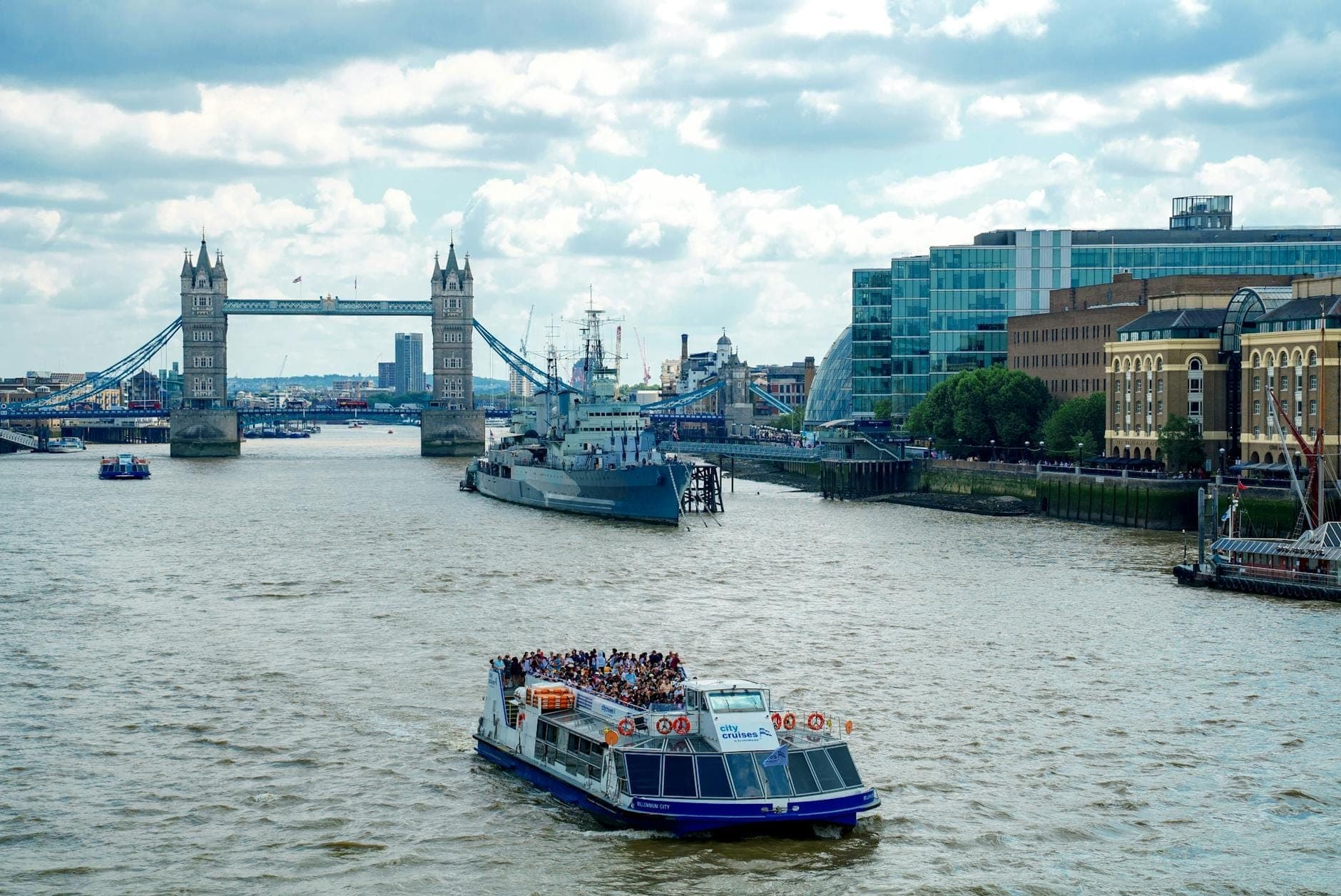 Sightseeing cruise boat full of passengers on the River Thames with Tower Bridge and modern London buildings in the background.