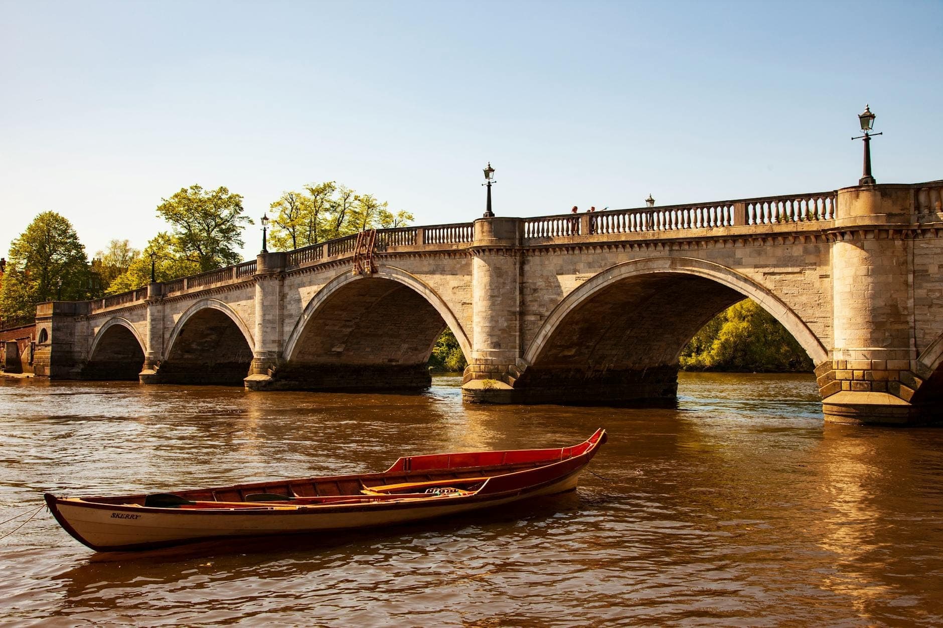A wooden rowing boat on the River Thames near a stone bridge in London under clear skies.