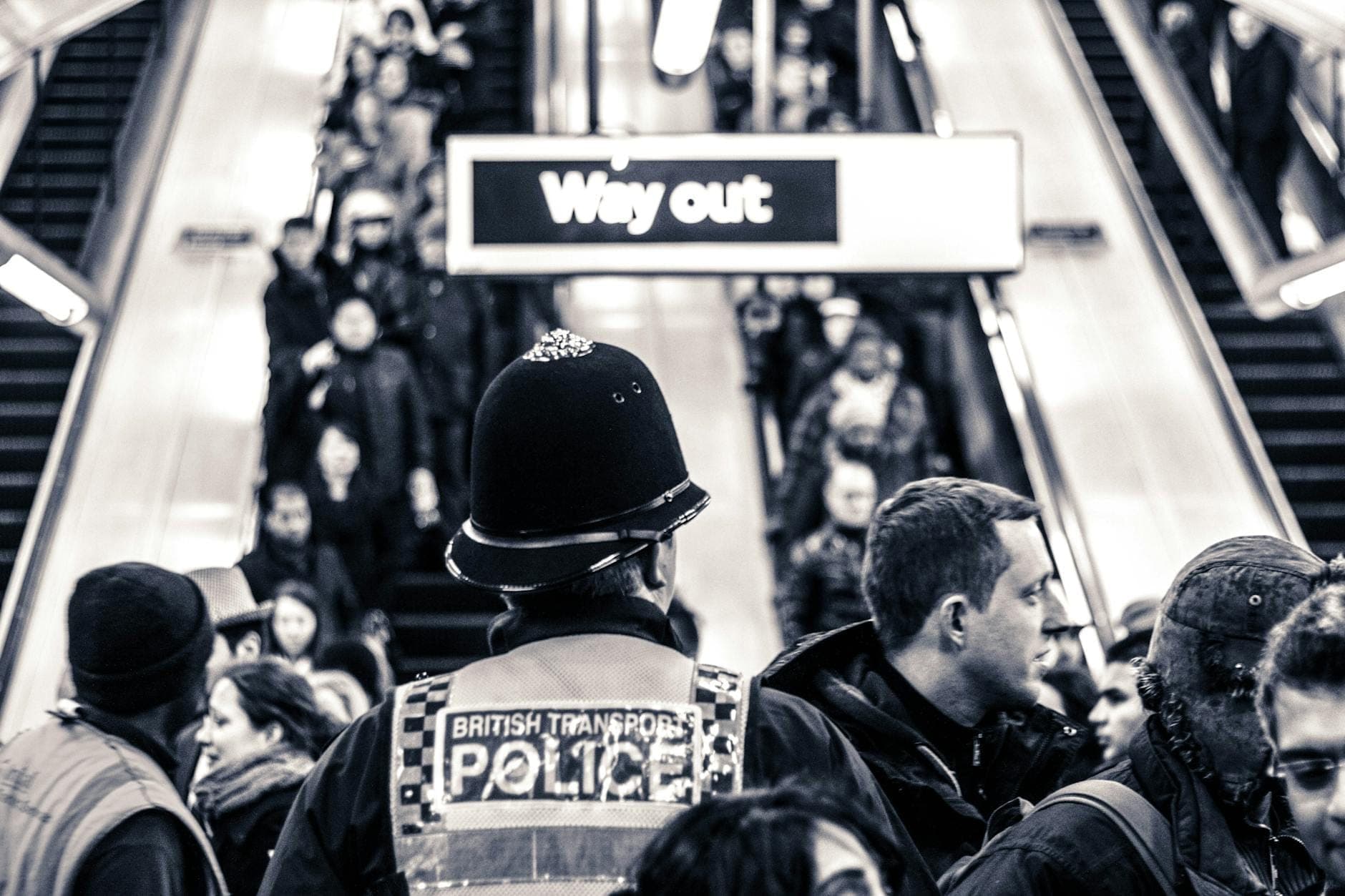 Un agent de la British Transport Police se tient au milieu d'une station de métro londonienne bondée, avec des voyageurs passant devant des escaliers mécaniques et un panneau 'Way out' au-dessus.