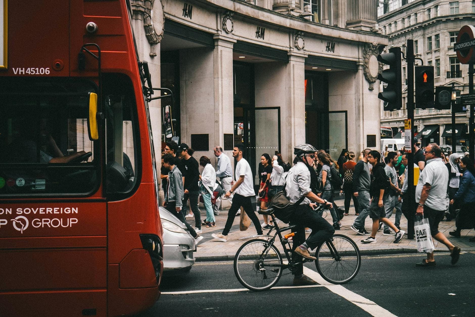 Scène de rue animée à Londres avec un bus rouge, un cycliste et des piétons traversant aux feux de circulation.
