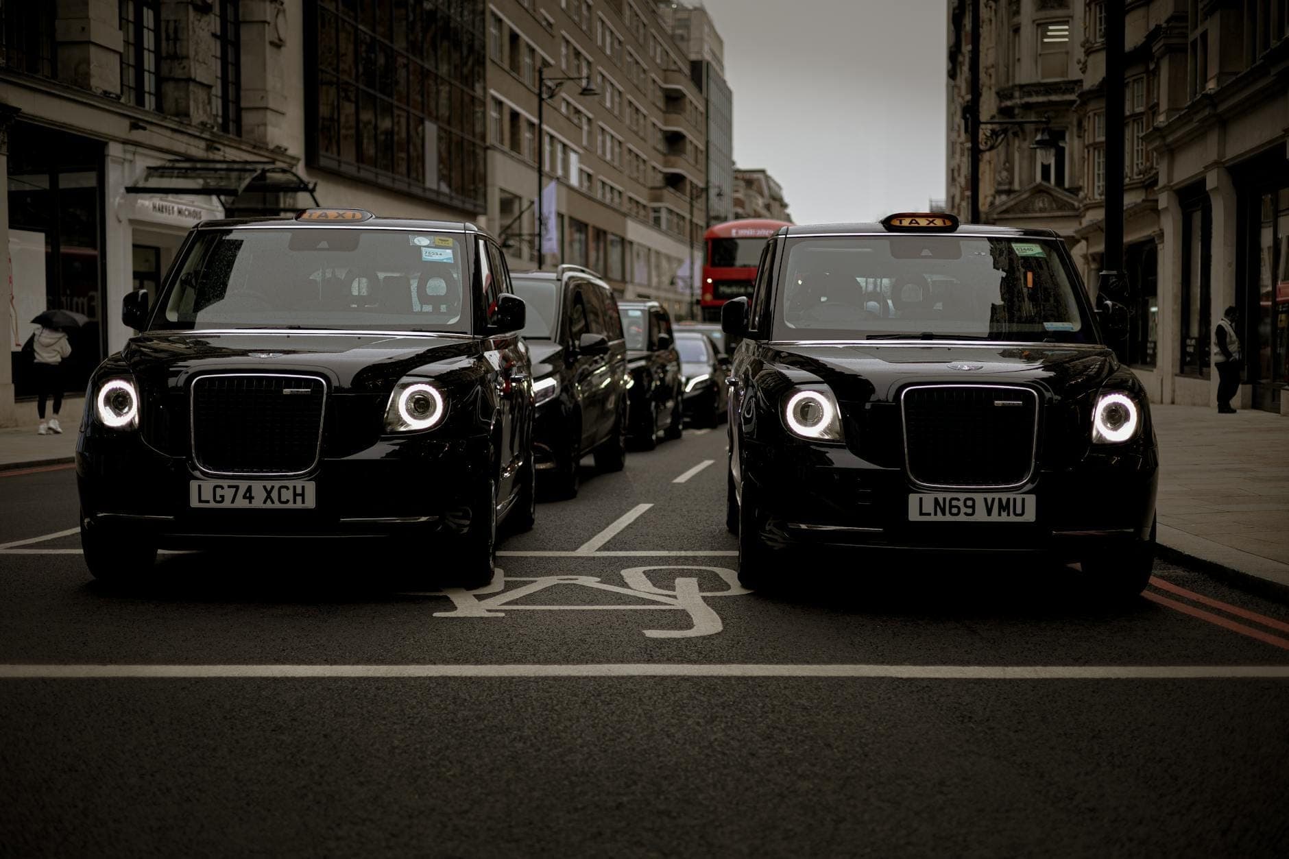 Deux taxis noirs emblématiques de Londres dans une rue animée avec d'autres véhicules et l'architecture classique de la ville visible.