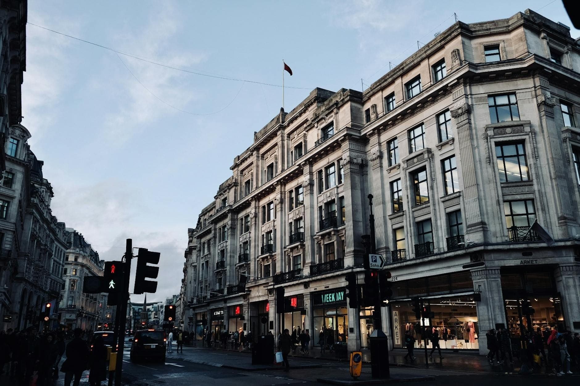 Concurrida calle comercial de Londres con grandes edificios históricos y tiendas en planta baja, personas caminando y farolas a la vista.