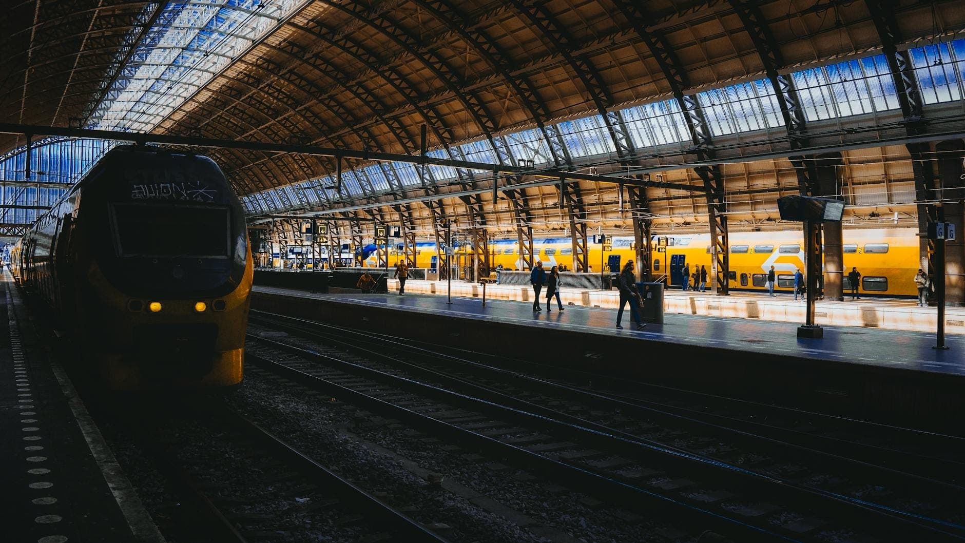 Trenes amarillos modernos en una amplia estación iluminada con techo de cristal arqueado y viajeros en el andén, evocando el trayecto entre grandes ciudades europeas.