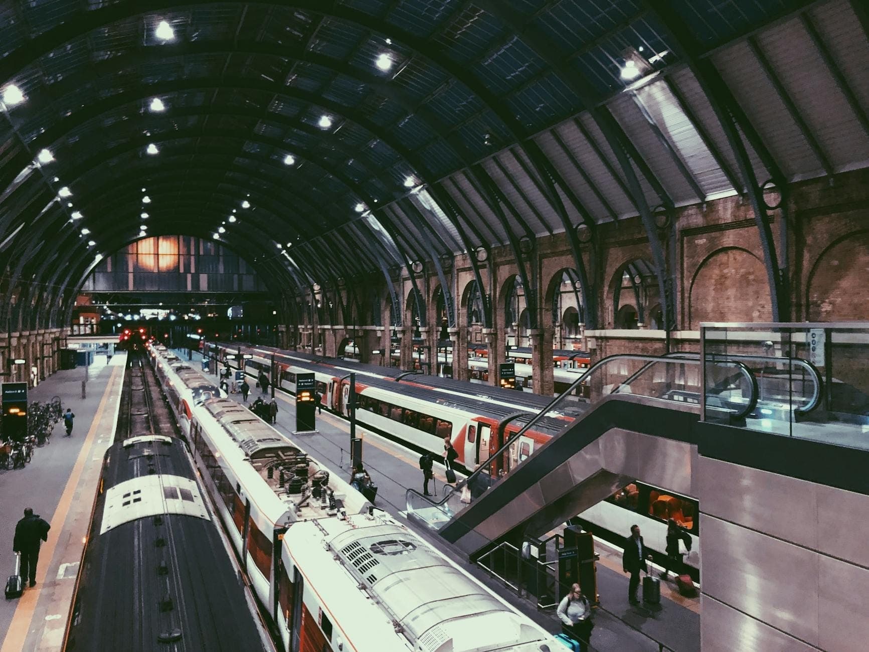 Interior movimentado de uma estação de trem com vários trens, teto de vidro arqueado e viajantes caminhando pelas plataformas em um terminal londrino agitado.