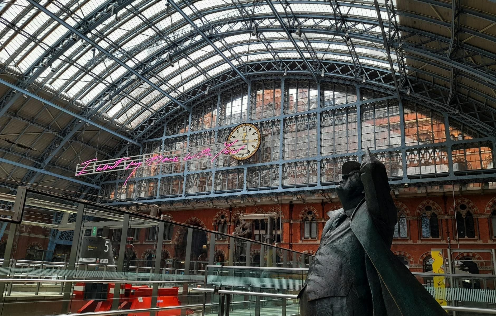 Interior da estação London St Pancras com teto de vidro arqueado, grande relógio de estação e estátua de um homem em primeiro plano.