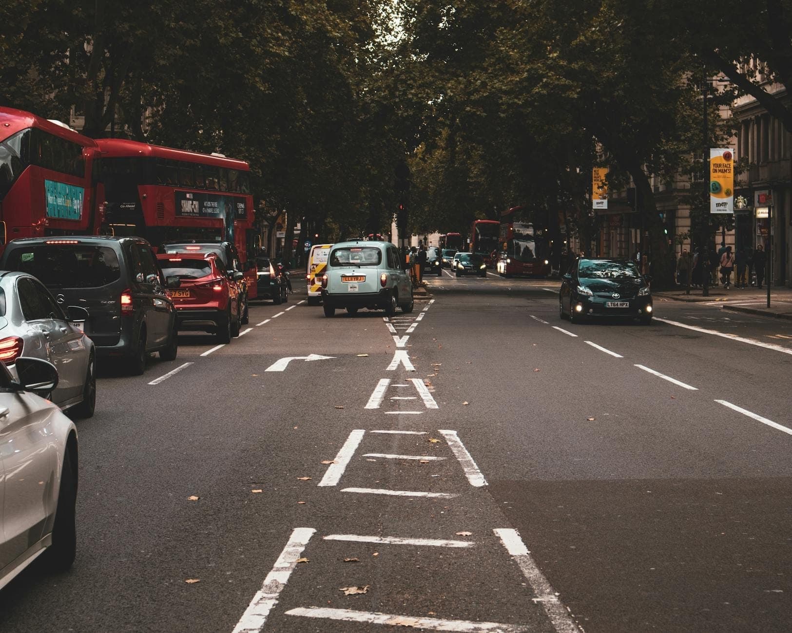 Trânsito típico de uma rua de Londres com carros, táxis pretos e ônibus vermelhos de dois andares sob árvores verdes