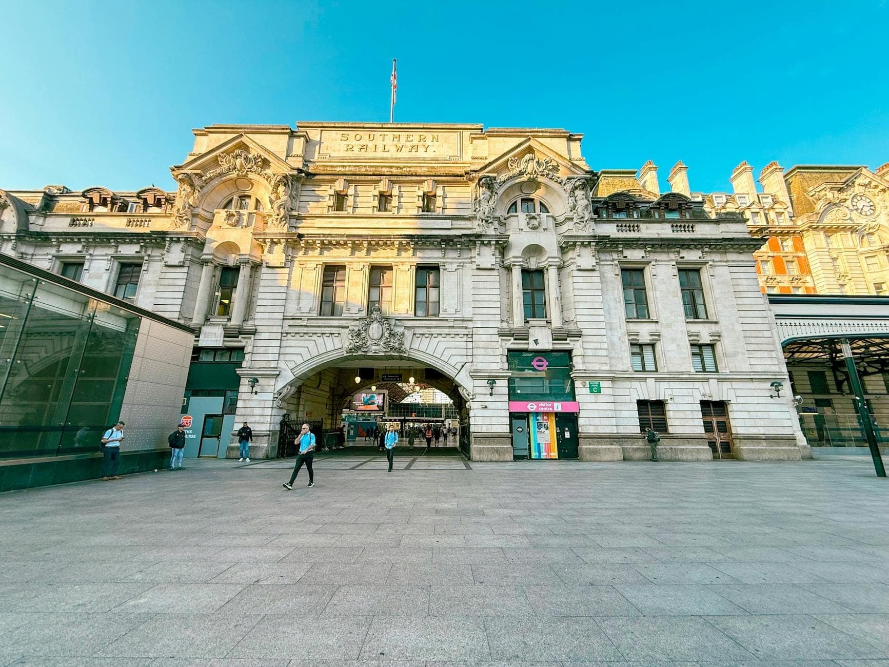 Szeroki widok na zewnątrz London Victoria Coach Station z ludźmi spacerującymi przed wejściem pod błękitnym niebem.