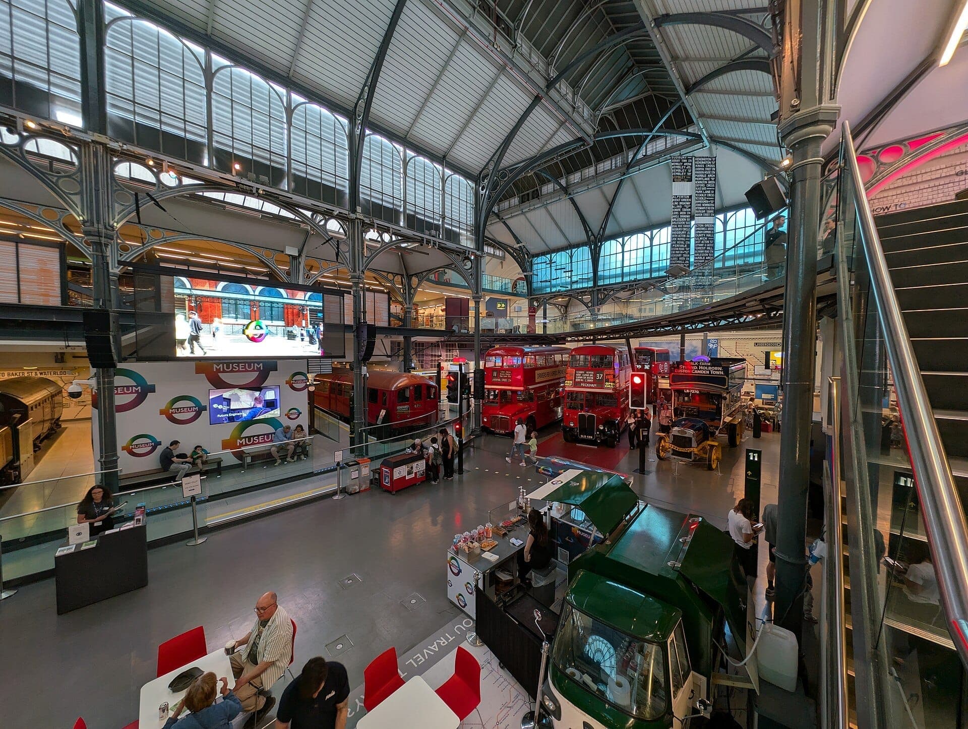 Innenansicht des London Transport Museum mit historischen roten Bussen, einem historischen U-Bahn-Wagen und viktorianischen Eisenkonstruktionen unter einem Glasdach.