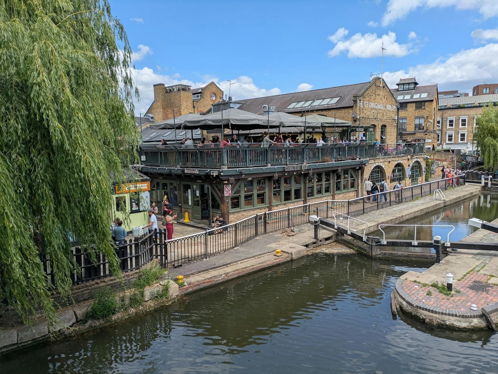 Eine lebhafte Kanalszene am Camden Market in London, mit Menschen, die am Wasser entlanggehen, historischen Gebäuden und einer Schleuse unter blauem Himmel.