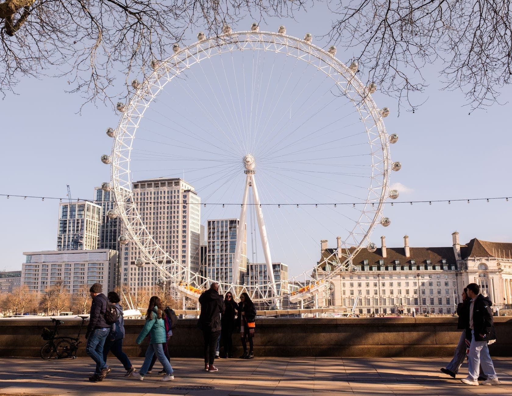Menschen, die am South Bank spazieren gehen, mit dem London Eye und Stadtgebäuden im Hintergrund an einem klaren Tag.