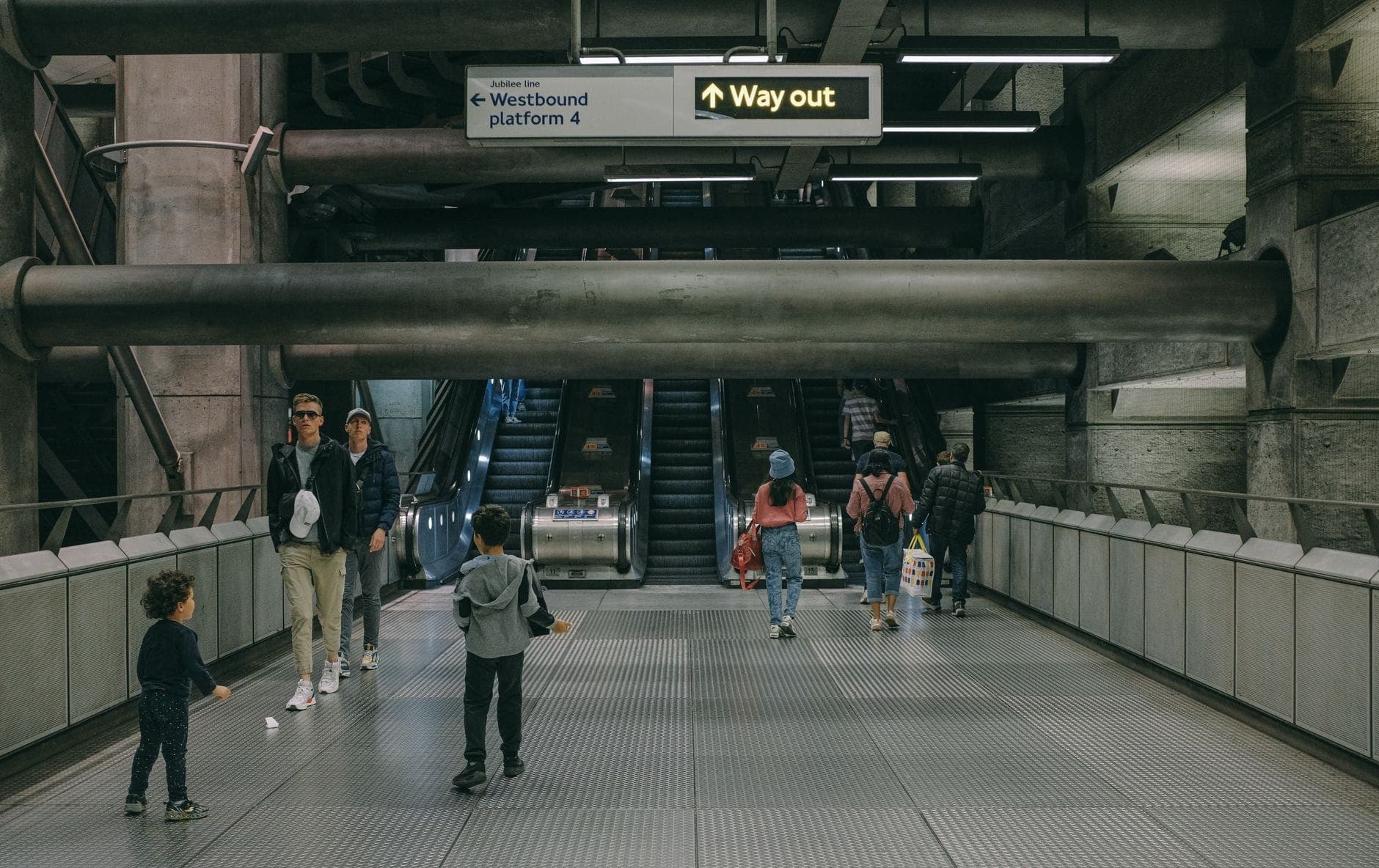 Famiglie con bambini che camminano all'interno di una stazione della London Underground con scale mobili e segnaletica 'Way out' in alto.