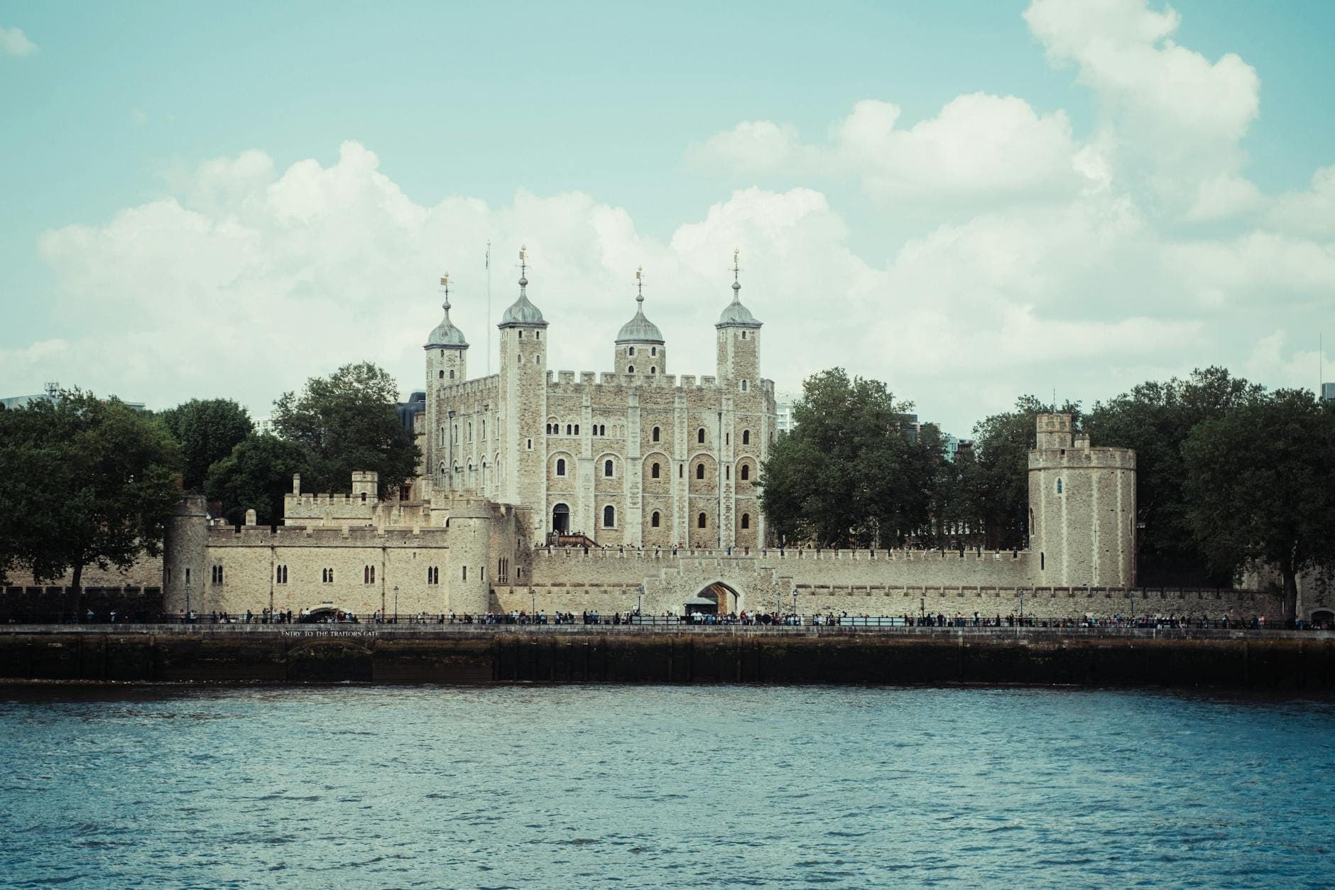 La Torre di Londra vista dall'altra sponda del fiume, con le sue iconiche mura in pietra e torrette su uno sfondo di cielo azzurro con nuvole.