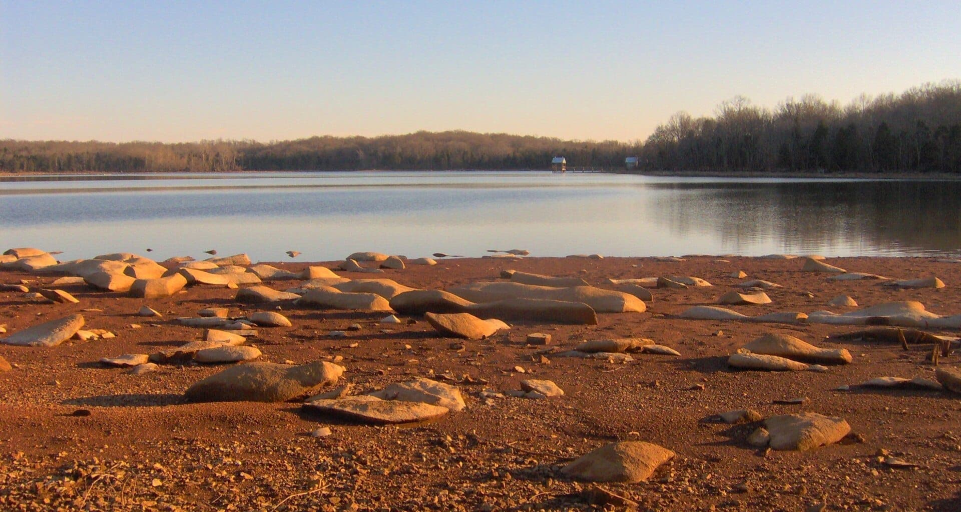 Rocky shoreline beside calm water of Couchville Lake at Long Hunter State Park with trees lining the distant horizon under a clear sky at sunset.