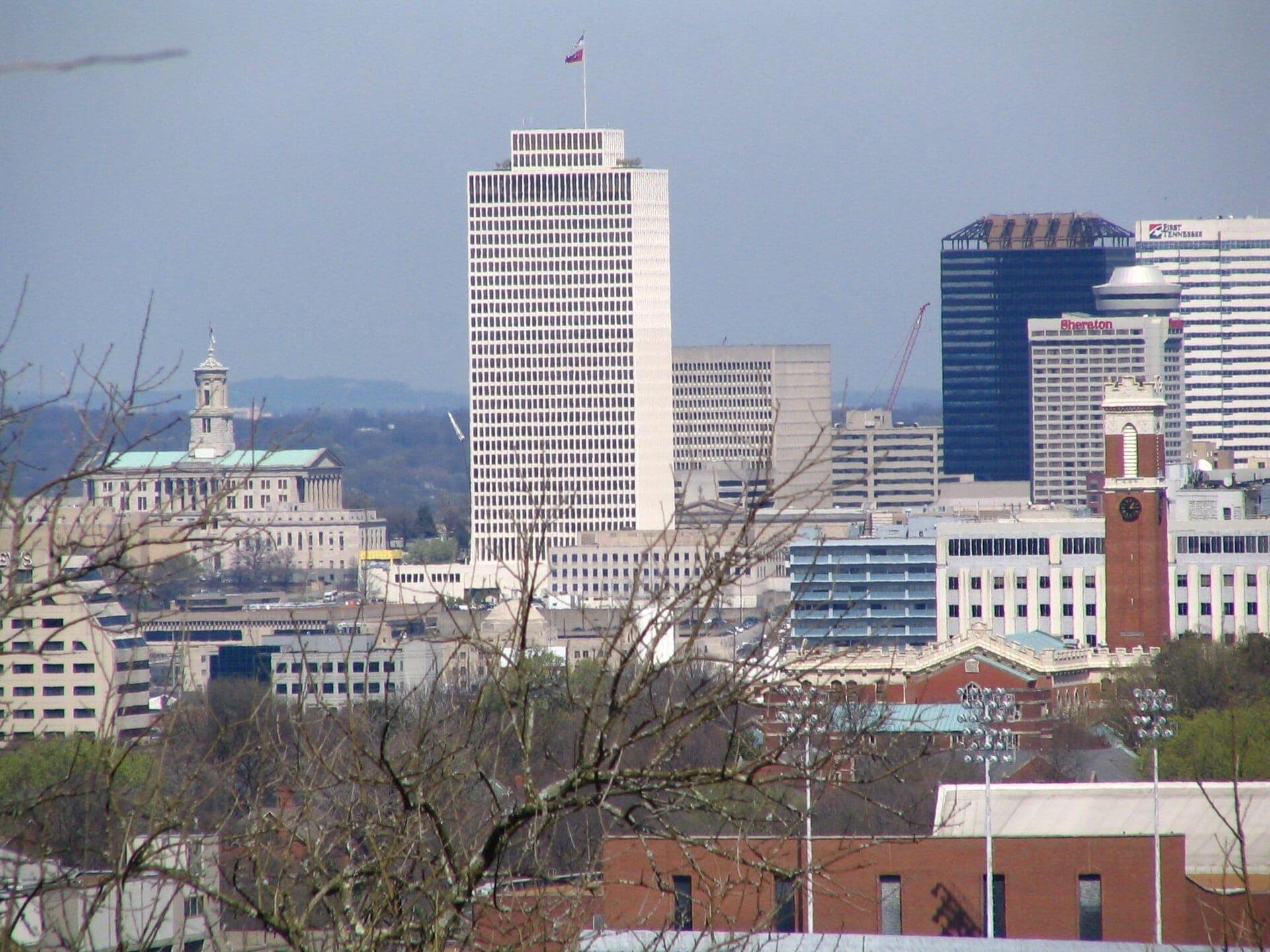 Horizonte del centro de Nashville visto desde Love Circle, con altos edificios de oficinas, el State Capitol, una torre con reloj y ramas de árboles en primer plano.