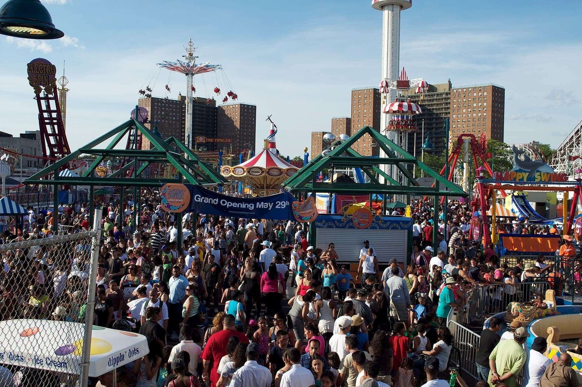 ฝูงชนจำนวนมากเต็มไปหมดที่ Luna Park บน Coney Island ในวันที่อากาศแจ่มใส ล้อมรอบด้วยเครื่องเล่นสีสันสดใส ป้ายแบนเนอร์ และอาคารในเมืองเป็นฉากหลัง