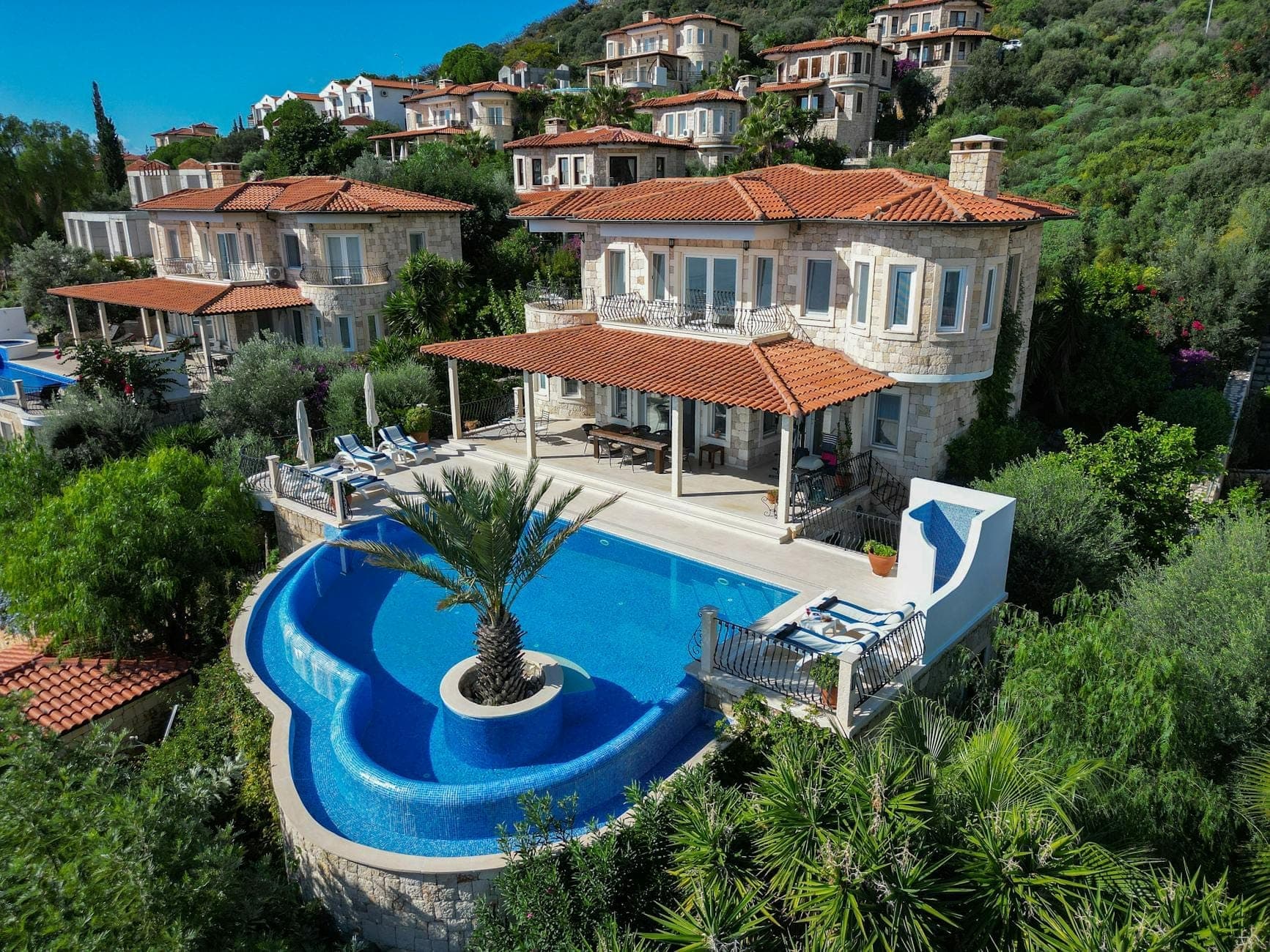 Luftaufnahme einer luxuriösen Steinvilla mit privatem Infinity-Pool, umgeben von üppigem Grün und anderen gehobenen Villen an einem Berghang