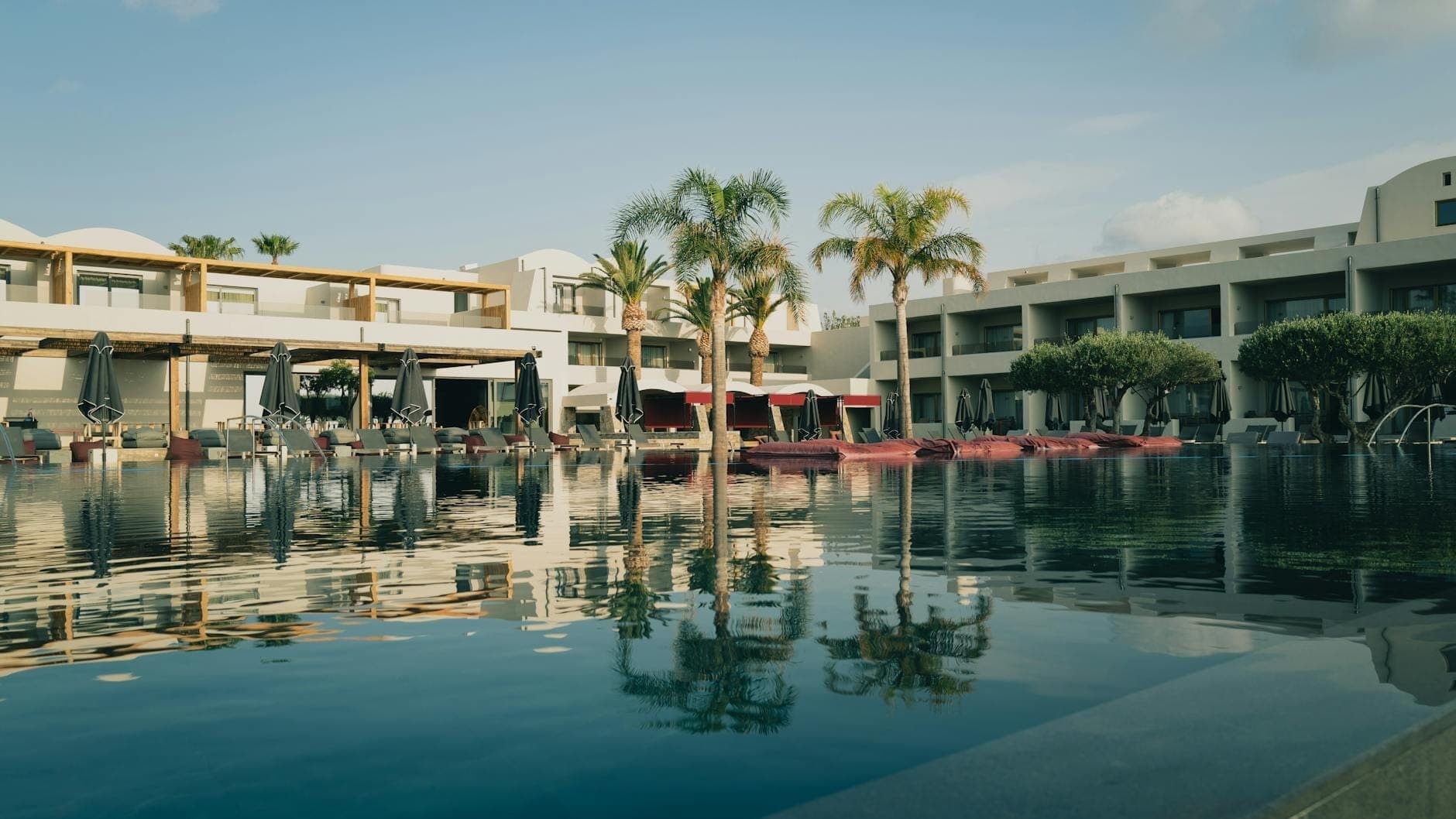 Luxusresort mit moderner Architektur, Palmen und Liegestühlen, die sich in einem ruhigen Pool unter blauem Himmel auf Kreta spiegeln.