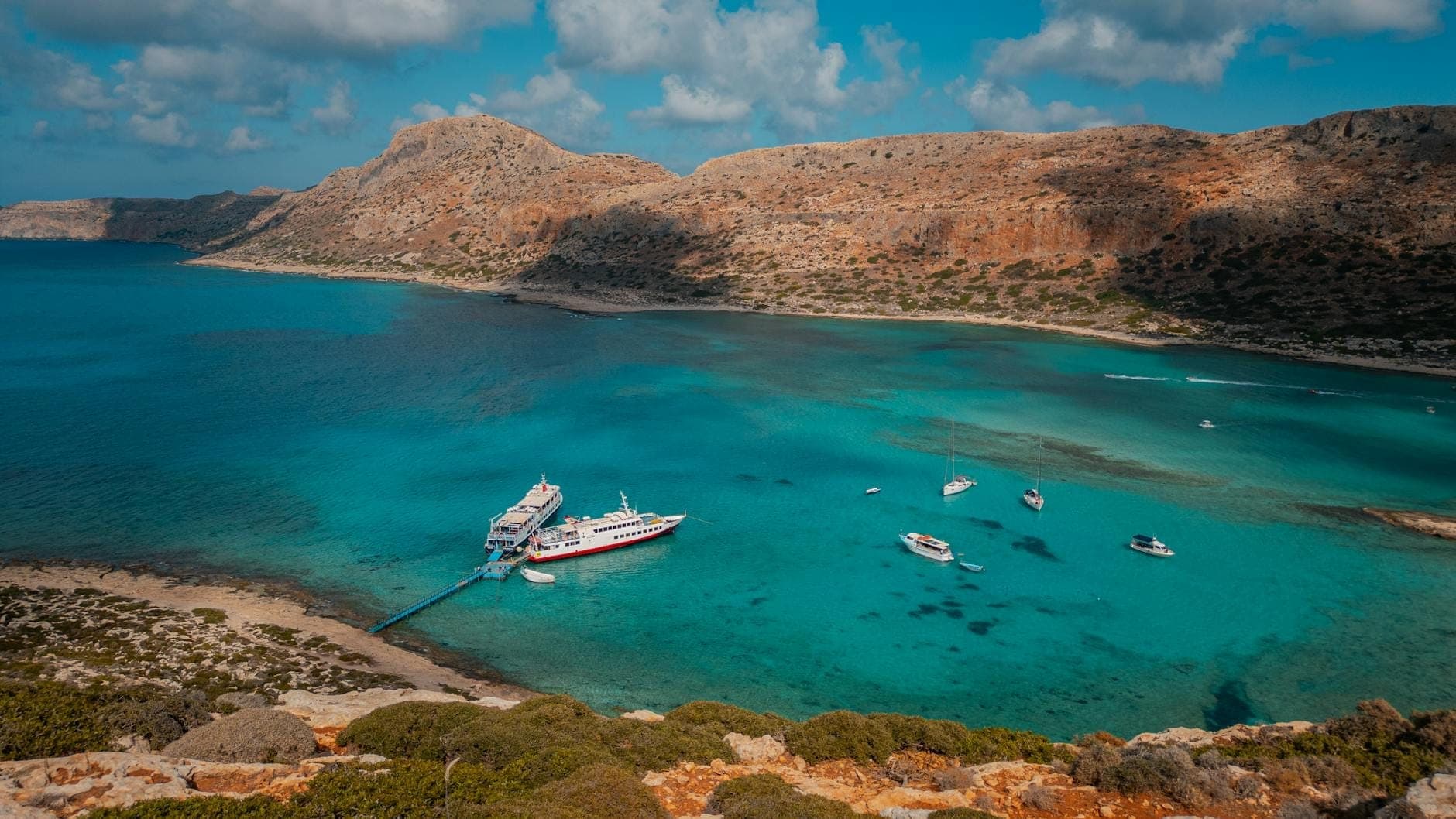 Türkisfarbene Bucht auf Kreta mit Luxusyachten und einem großen Boot, das in der Nähe einer felsigen Küste unter blauem Himmel angelegt hat.