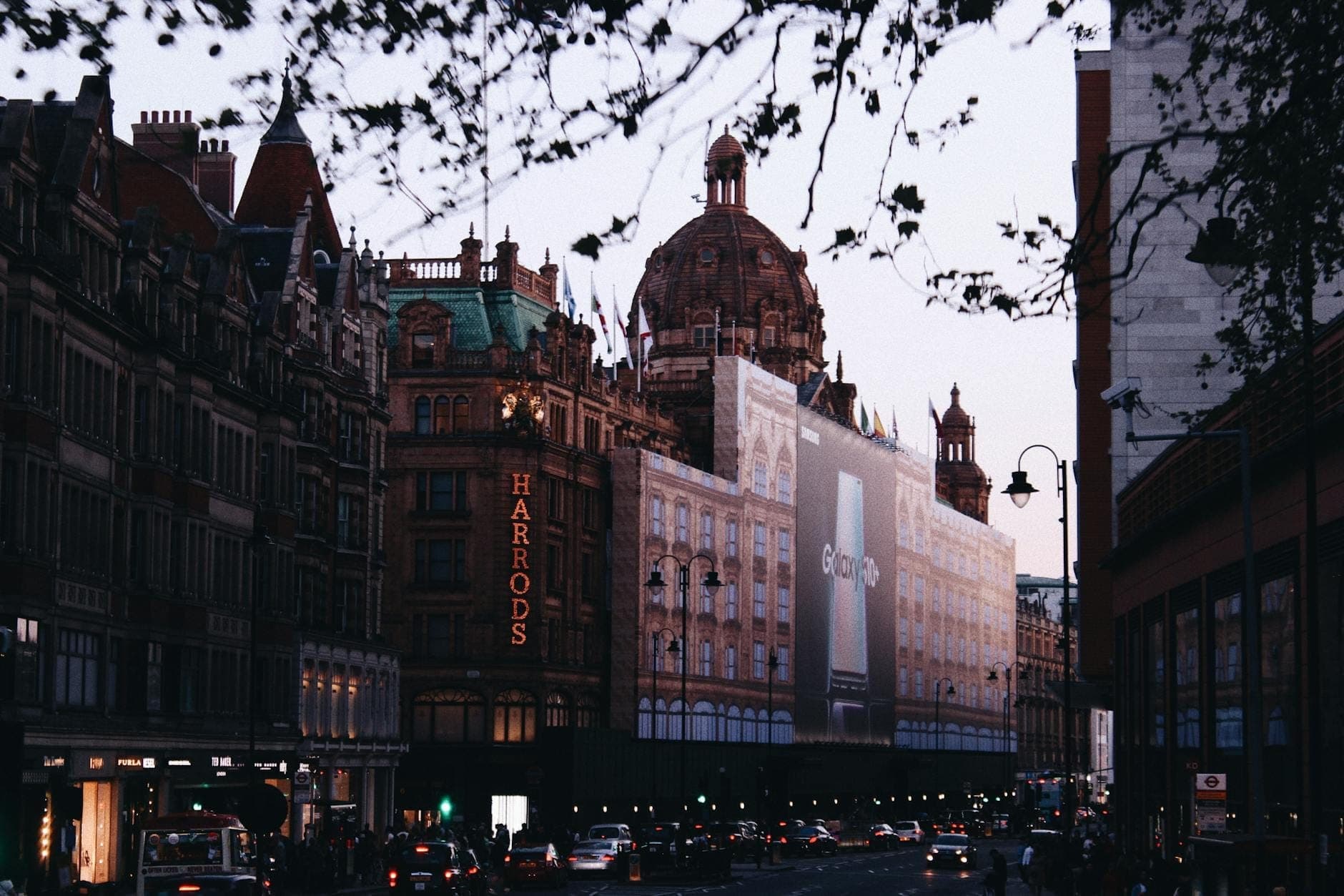 Londra'daki lüks alışveriş merkezi Harrods'un alacakaranlıkta klasik mimarisi ve önündeki yoğun trafikle sokak görünümü.