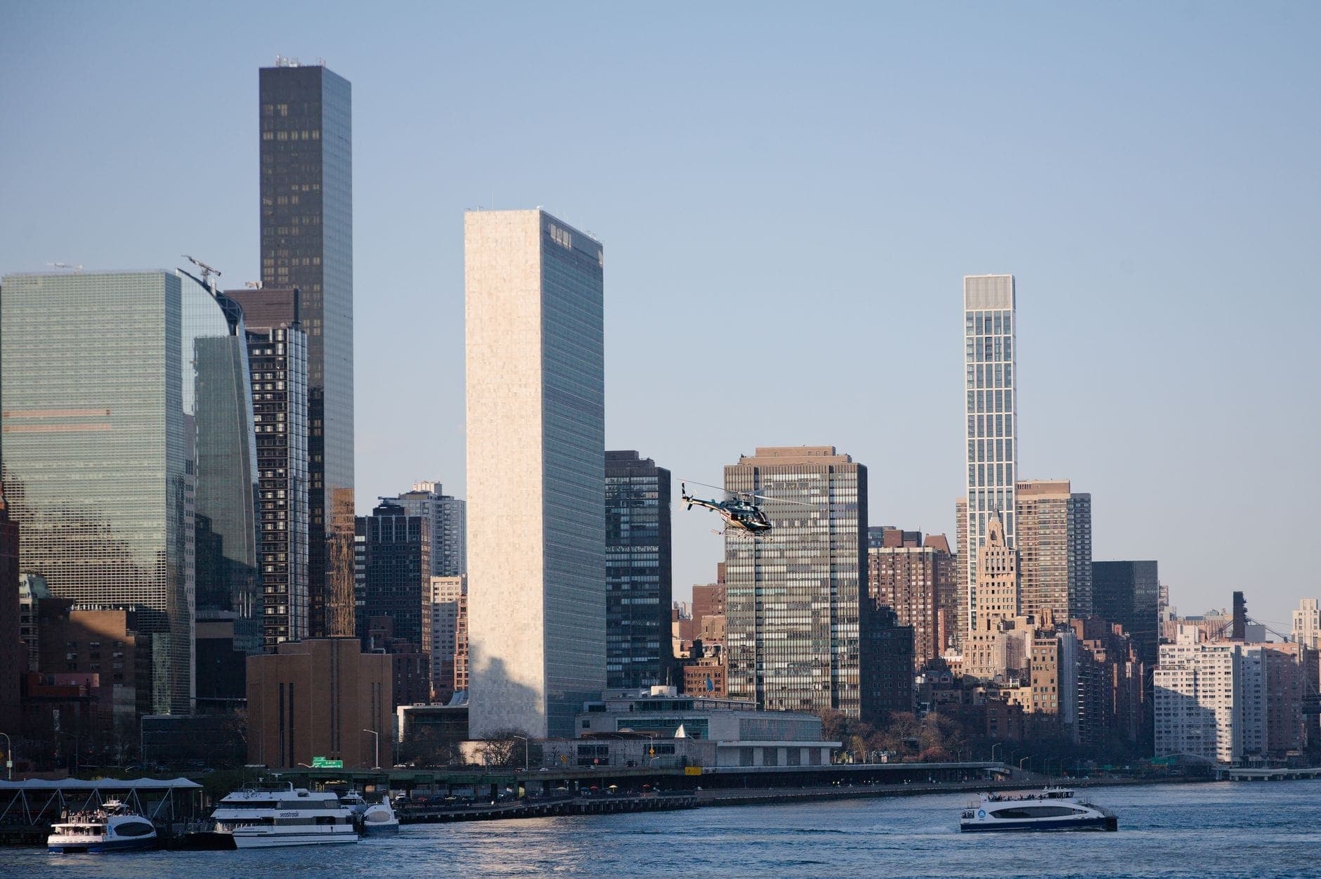 เฮลิคอปเตอร์บินอยู่เหนือ East River พร้อมตึกระฟ้าของ New York City และเรือยอทช์บนผืนน้ำในเวลากลางวัน