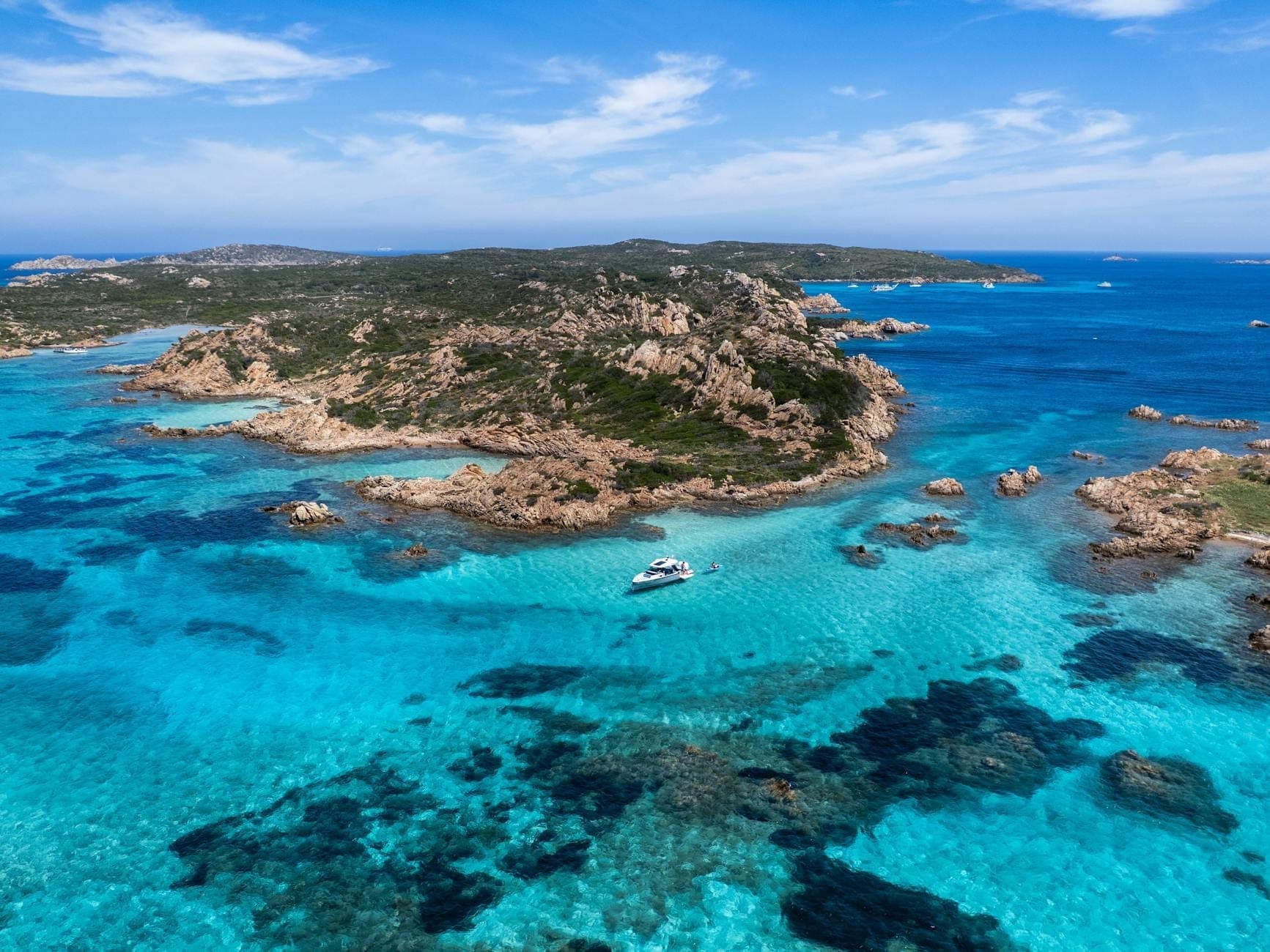 Vista aérea de aguas turquesa, costa escarpada y un yate anclado cerca de las pintorescas orillas rocosas de Cerdeña bajo cielos azules.