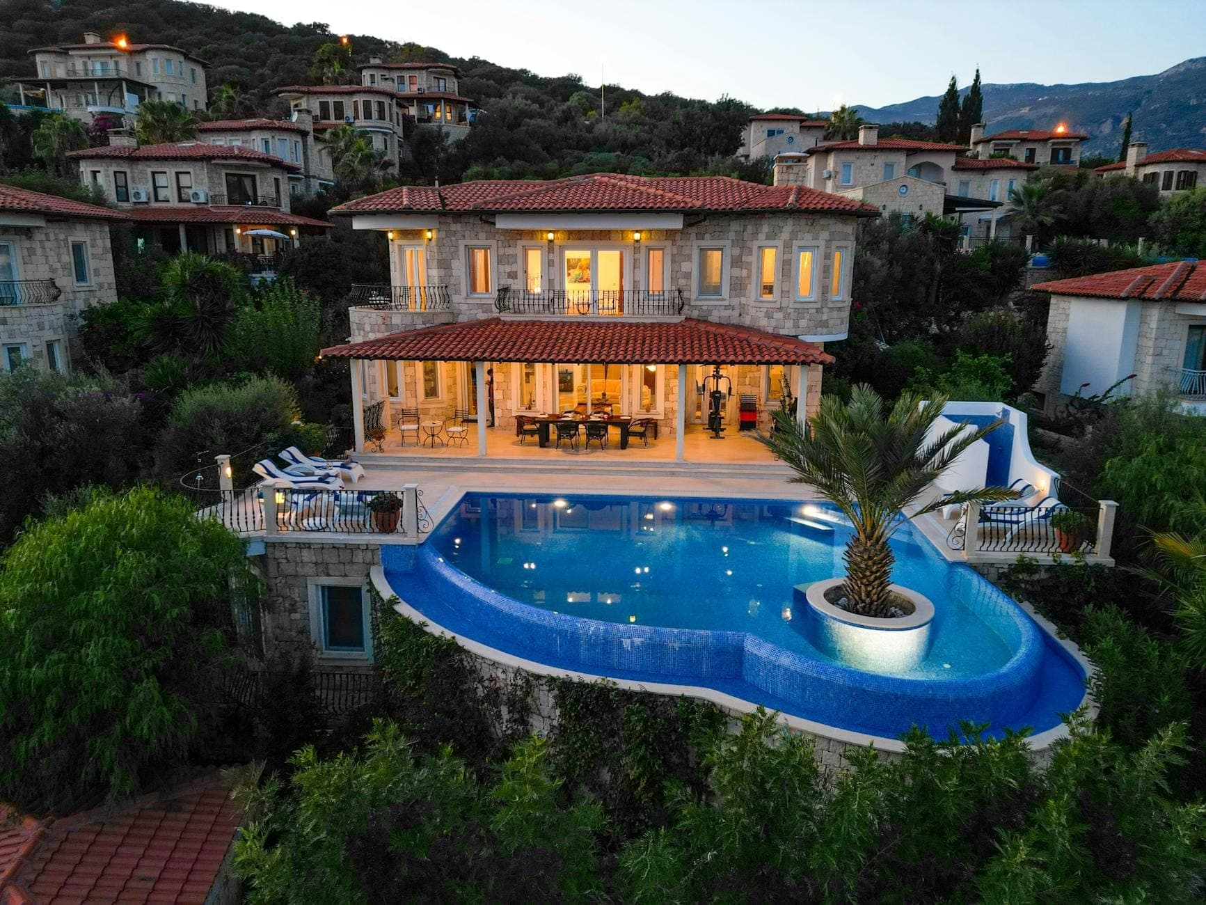 Una villa grande y lujosa con piscina infinita privada, rodeada de vegetación y casas en la ladera, al atardecer o al anochecer.