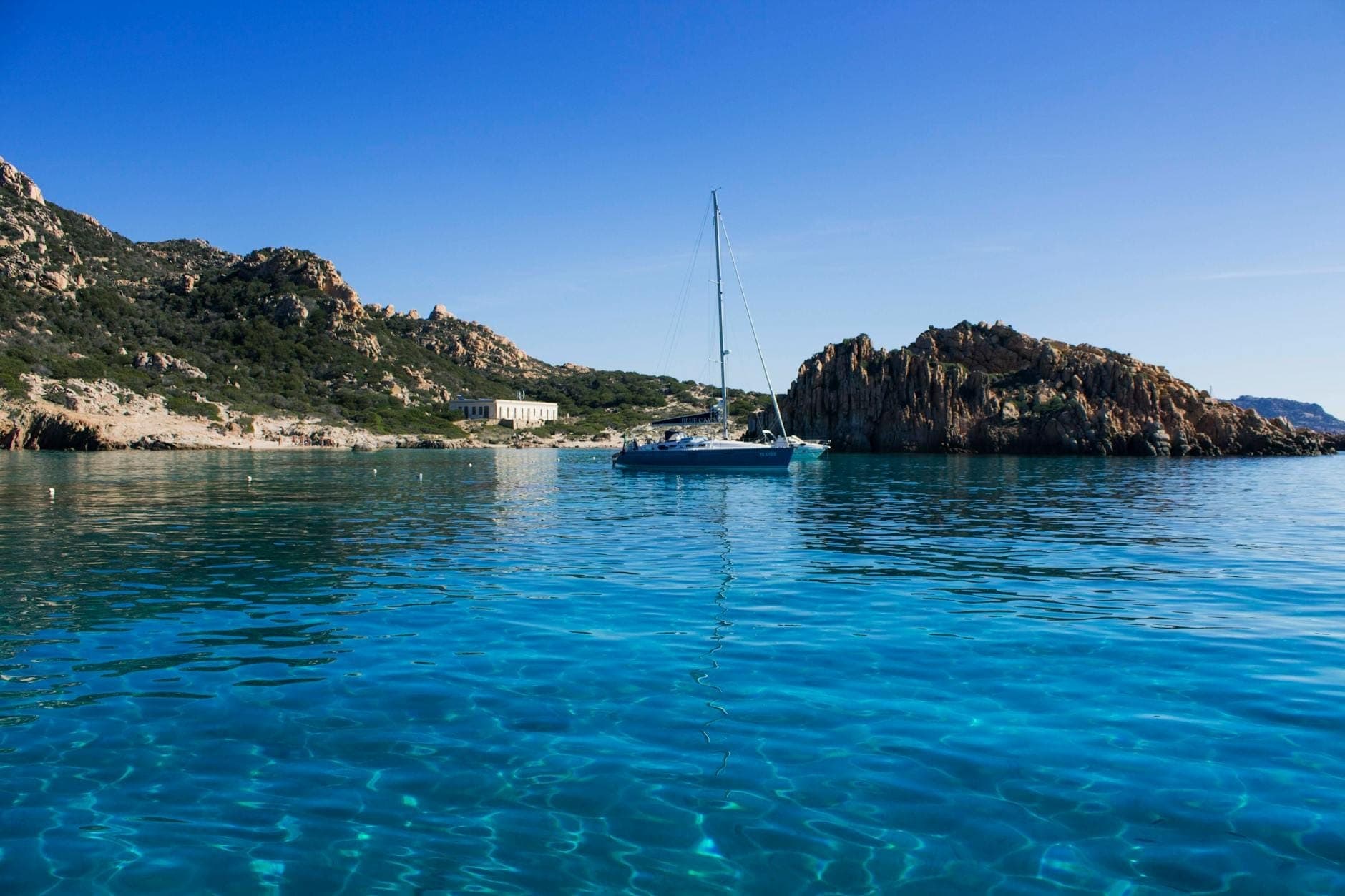 Velero anclado en aguas turquesas cristalinas rodeado por la rocosa costa de Cerdeña bajo un cielo azul brillante.