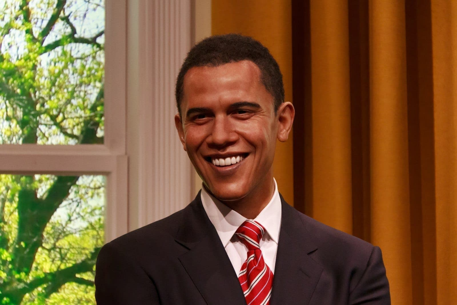 Wachsfigur des lächelnden Barack Obama bei Madame Tussauds London, mit goldenen Vorhängen und einem Fenster hinter ihm.